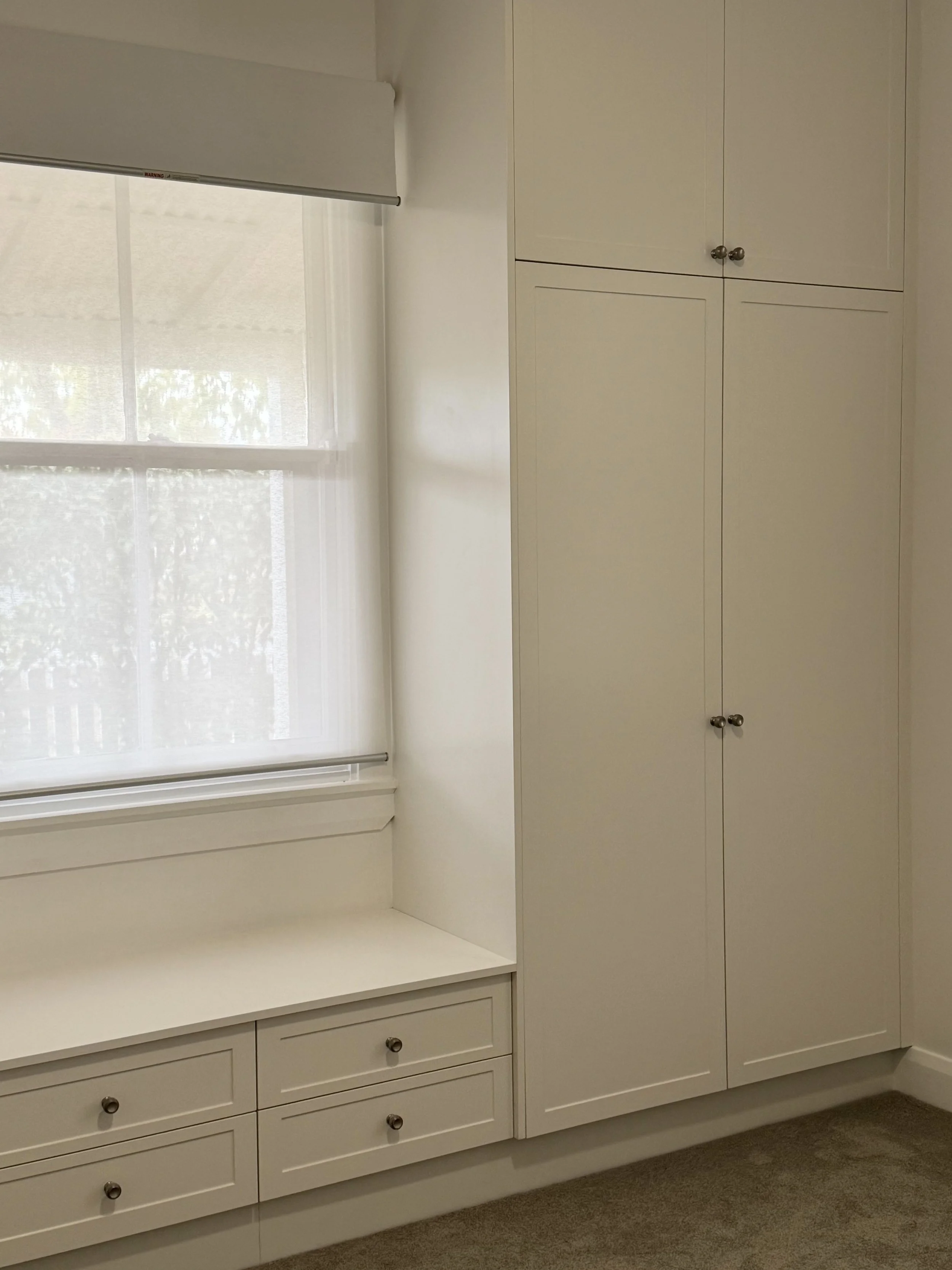 Interior of a room showing a large window with white blinds, a built-in white cabinet with multiple doors, drawers, and a window seat.