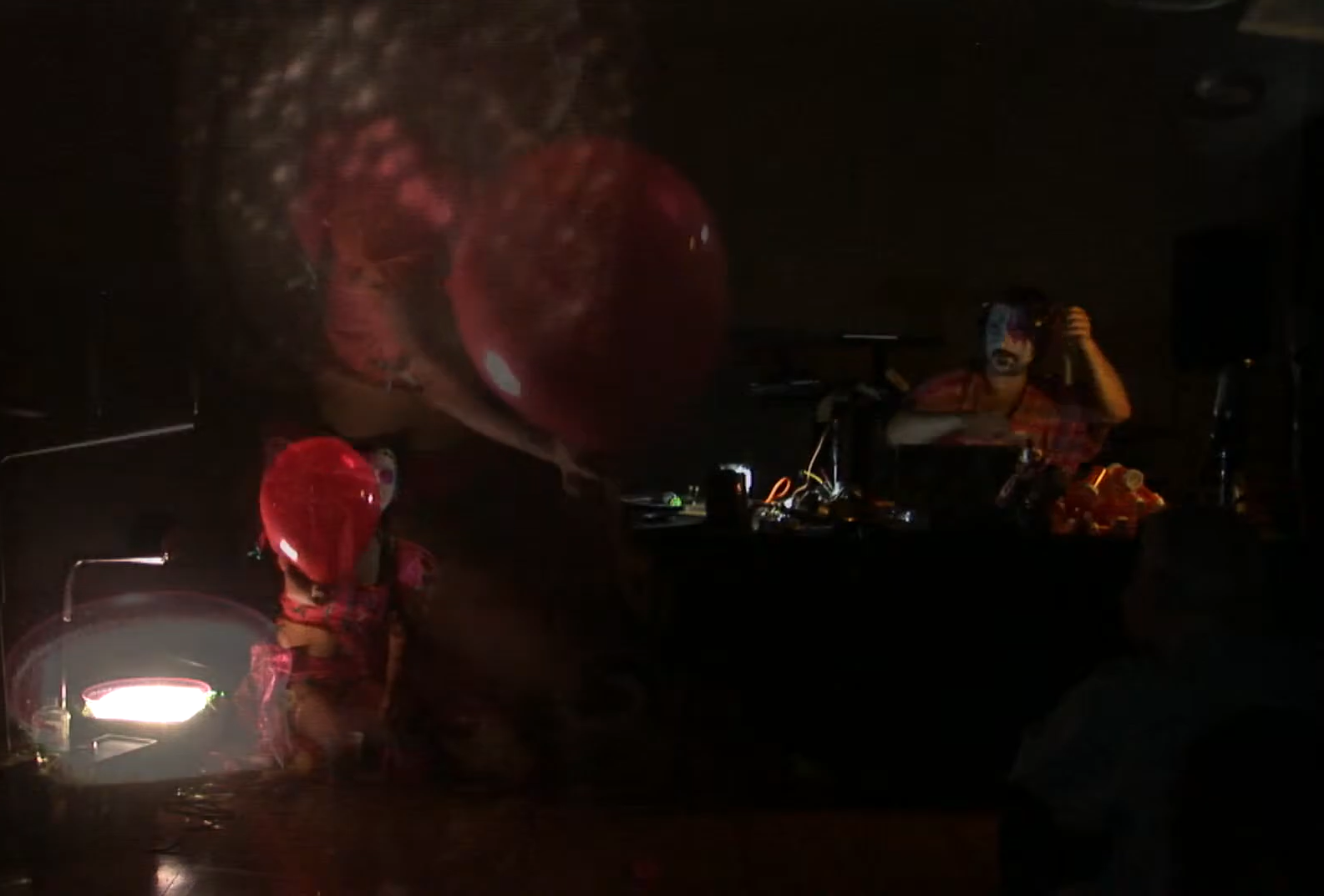 A person with face paint or makeup sitting at a table in a dark room, with a large red object above them and various items scattered on the table, illuminated by a small light.