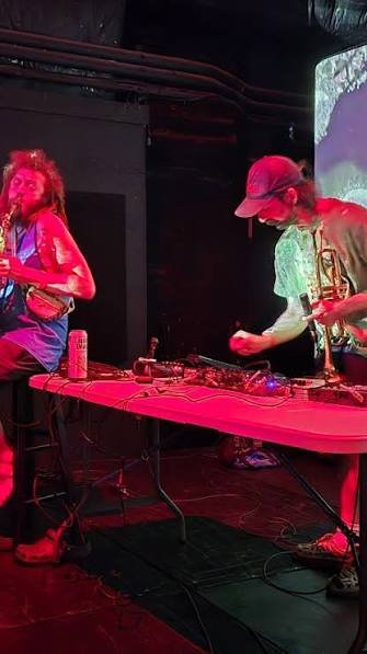 Two musicians performing on stage at a nightclub, one playing a saxophone and the other using DJ equipment.