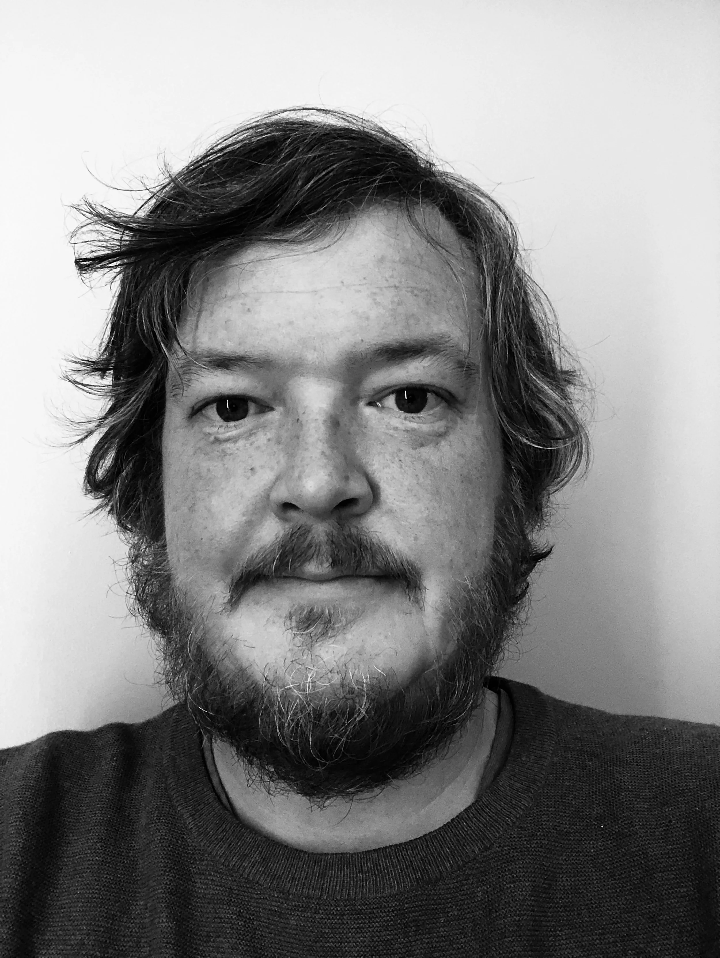 Close-up black and white photo of a man with curly hair, a beard, and a mustache, wearing a dark sweater, against a plain light-colored background.