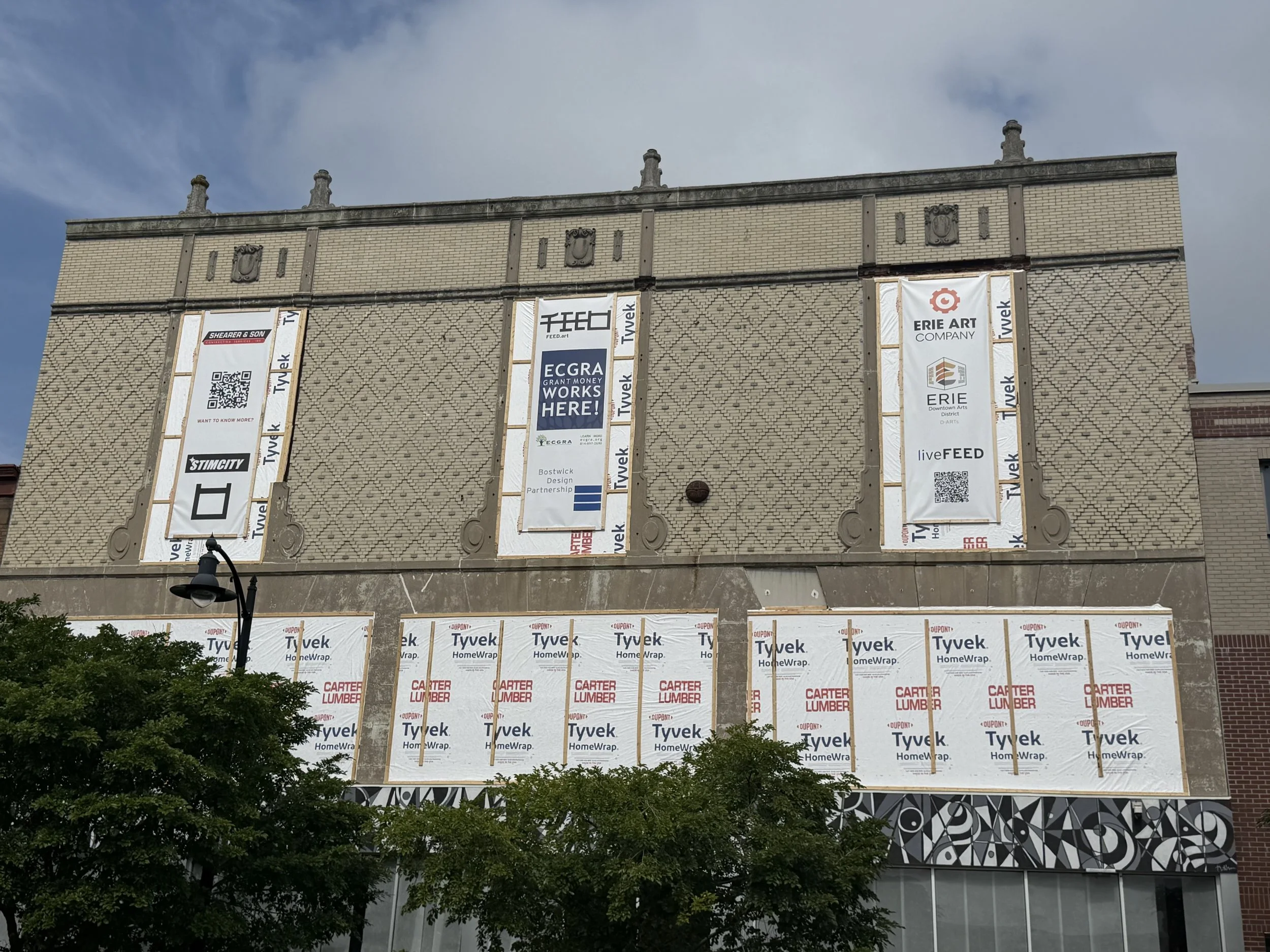 The image shows the side of a building under construction with scaffolding and building materials, including banners with brand names like Tyvek and Carter Lumber, and a part of the mural on the lower right corner.