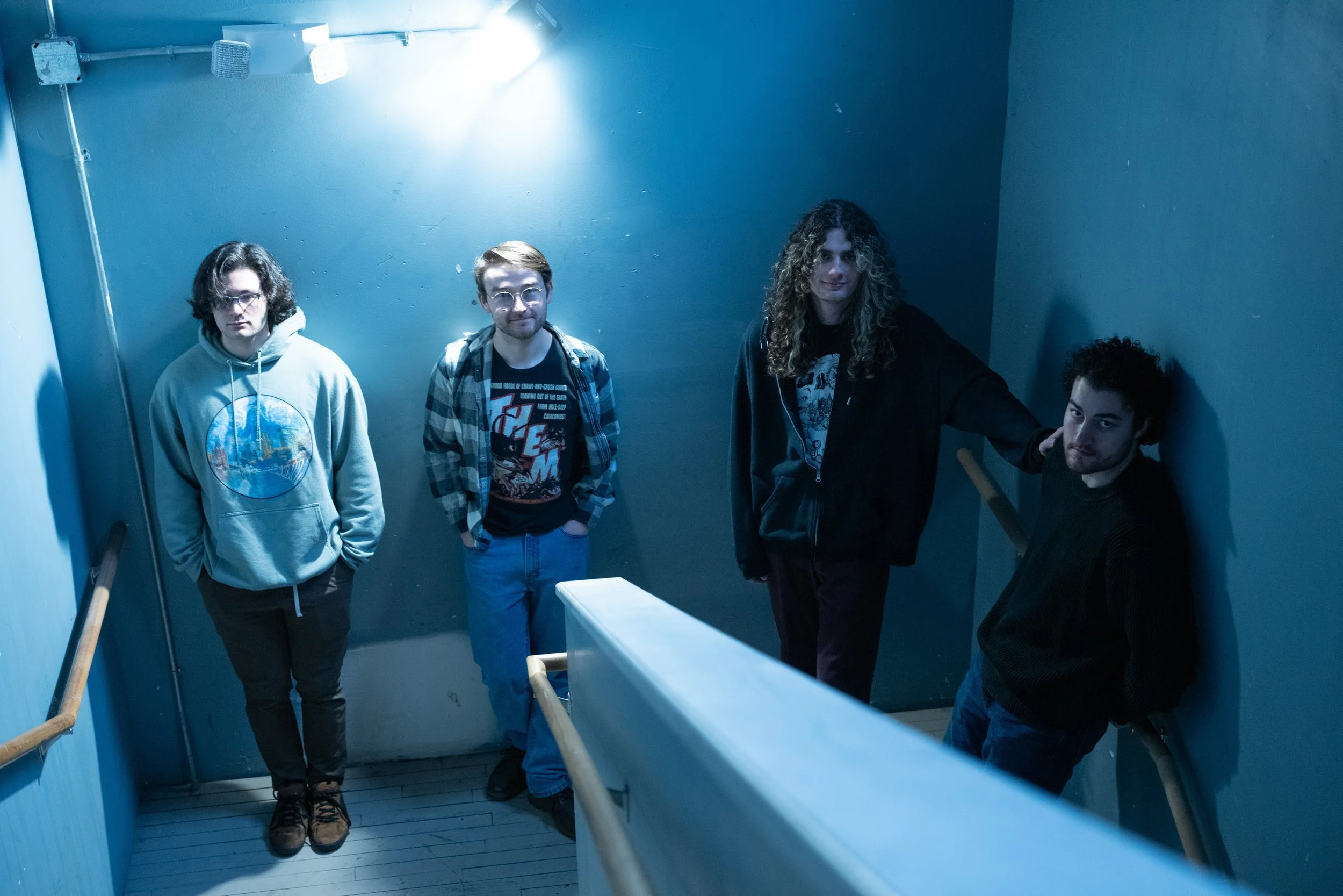 Four young men standing on a staircase inside a building, illuminated by blue-tinted lighting. They are casually dressed, with one wearing glasses, and are looking at the camera.