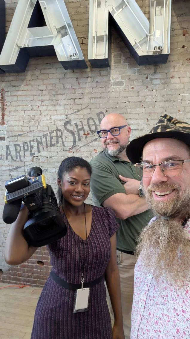 Three people smiling for a photo inside a building with a brick wall background. The woman on the left holds a camera and wears a patterned dress. The man in the middle has glasses and a beard, and the man on the right, also with glasses and a hat, t