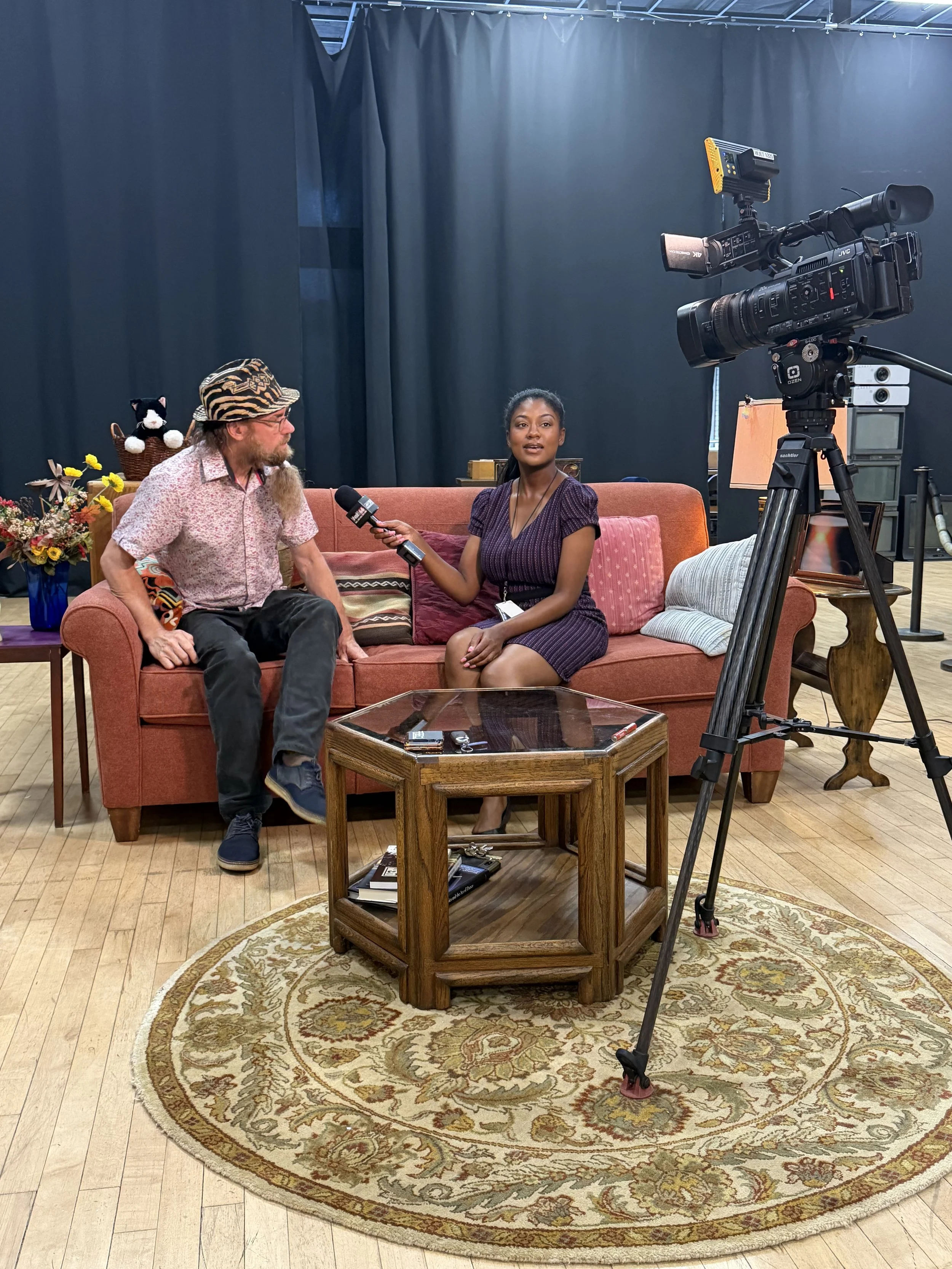 A woman interviewing a man on a sofa in a room with black curtains and camera equipment.