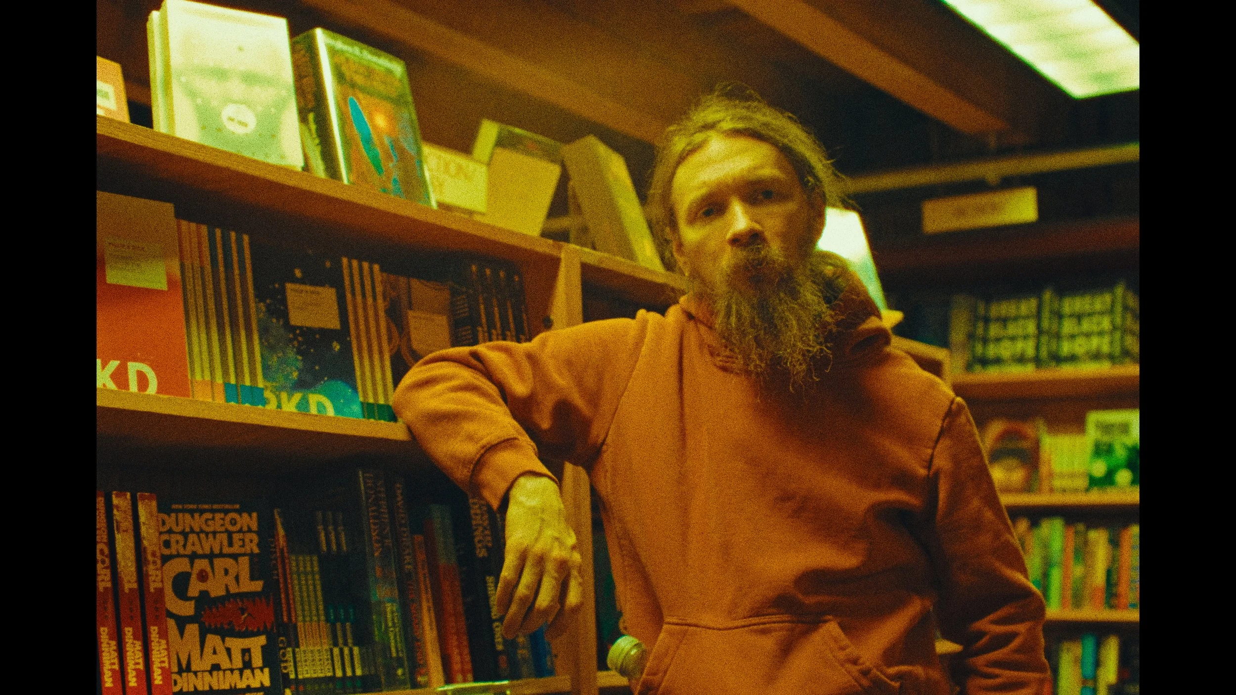A man with a beard and long hair standing in a bookstore, leaning against a shelf filled with colorful books.