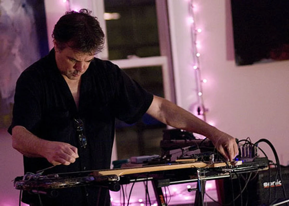 A man with dark hair and a black shirt is operating electronic music equipment in a room decorated with pink string lights.