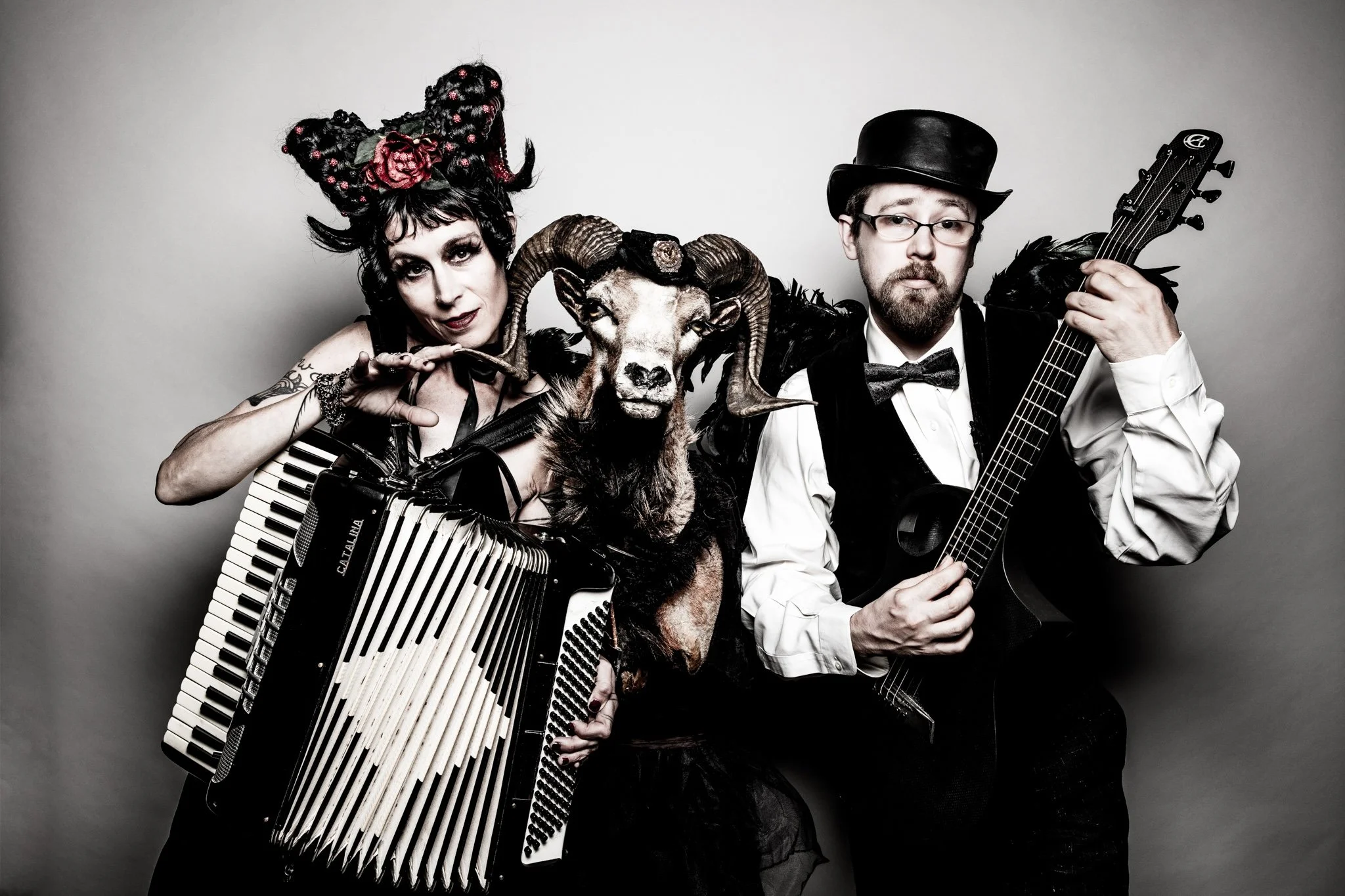 Two musicians, one woman with elaborate headpiece and an accordion, and one man with a hat and glasses holding a guitar, posing together against a plain background.