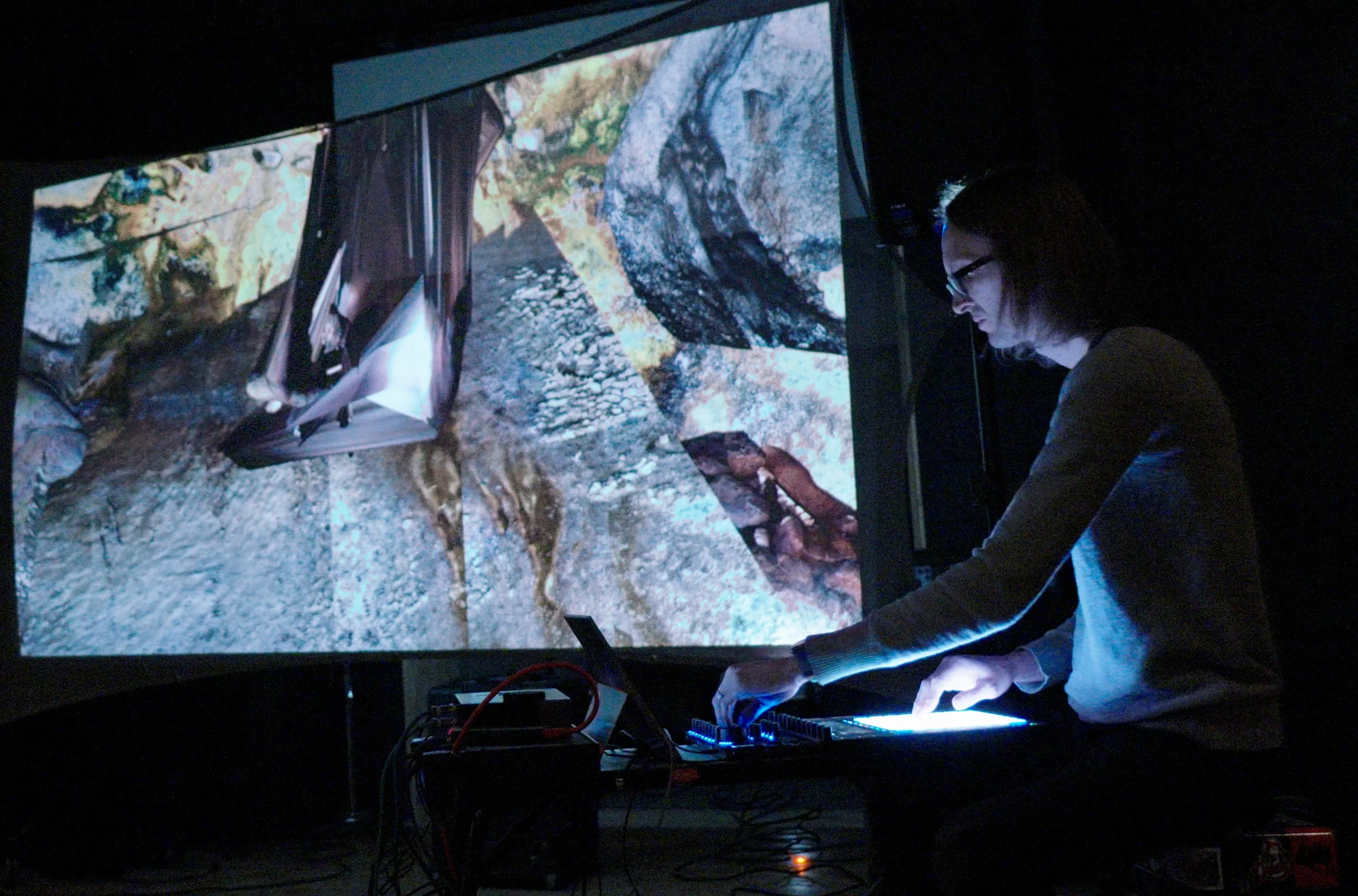 A person with glasses working with electronic equipment on a dark background, with a large projection of a rocky mountain landscape on the wall.