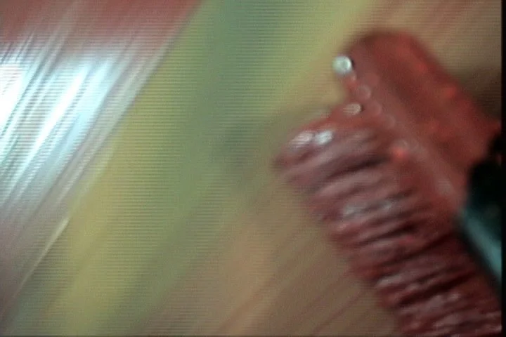 Blurred image showing a partial view of a person's face with glasses, wooden furniture, and a wall in the background.