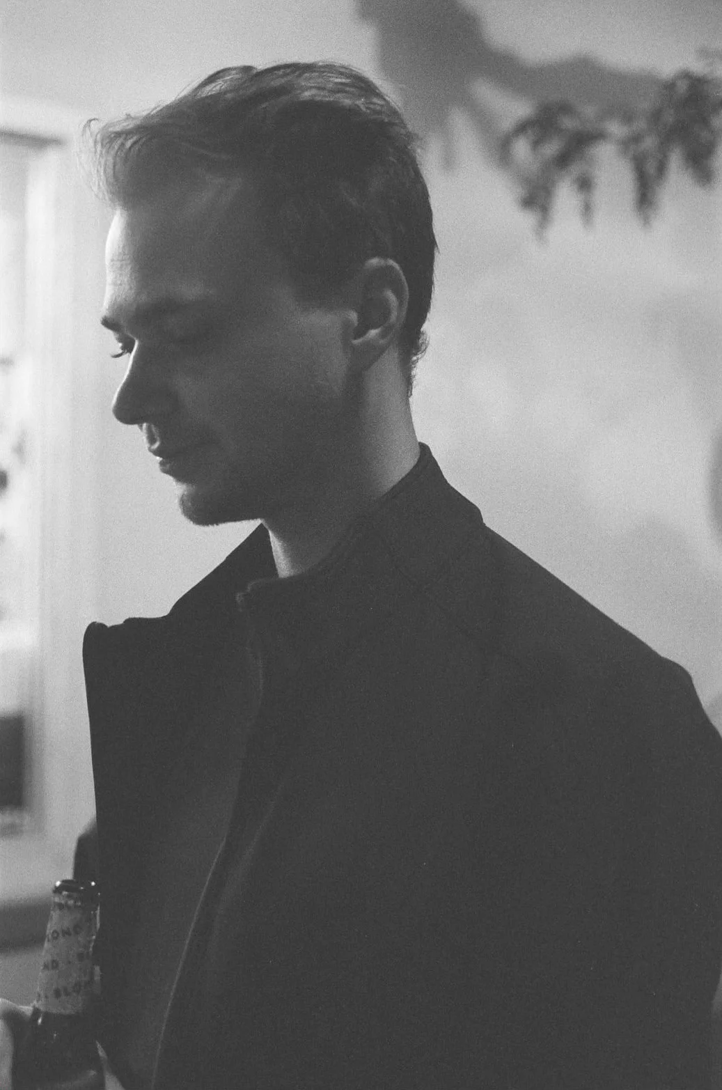 Black and white photo of a young man with short hair, looking down and holding a bottle, in an indoor setting with a blurred background.