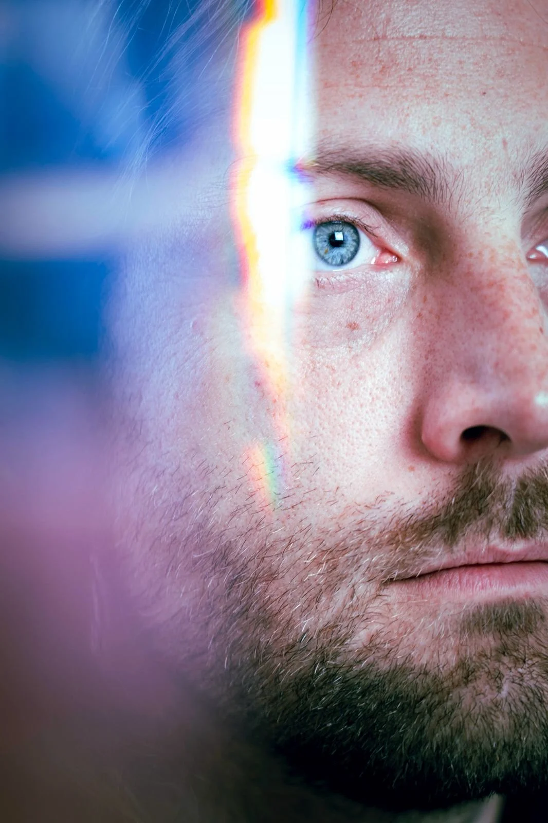 Close-up of a man's face with a blue eye, stubble, and a rainbow reflection on his cheek.