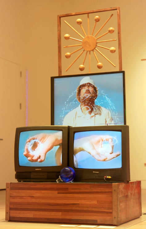 Three vintage televisions stacked on a wooden box, displaying visuals of hands holding spherical objects, and a person with water splashing on their face on the screens. A framed decorative wall piece with a gold geometric sunburst design is hanging in the background.