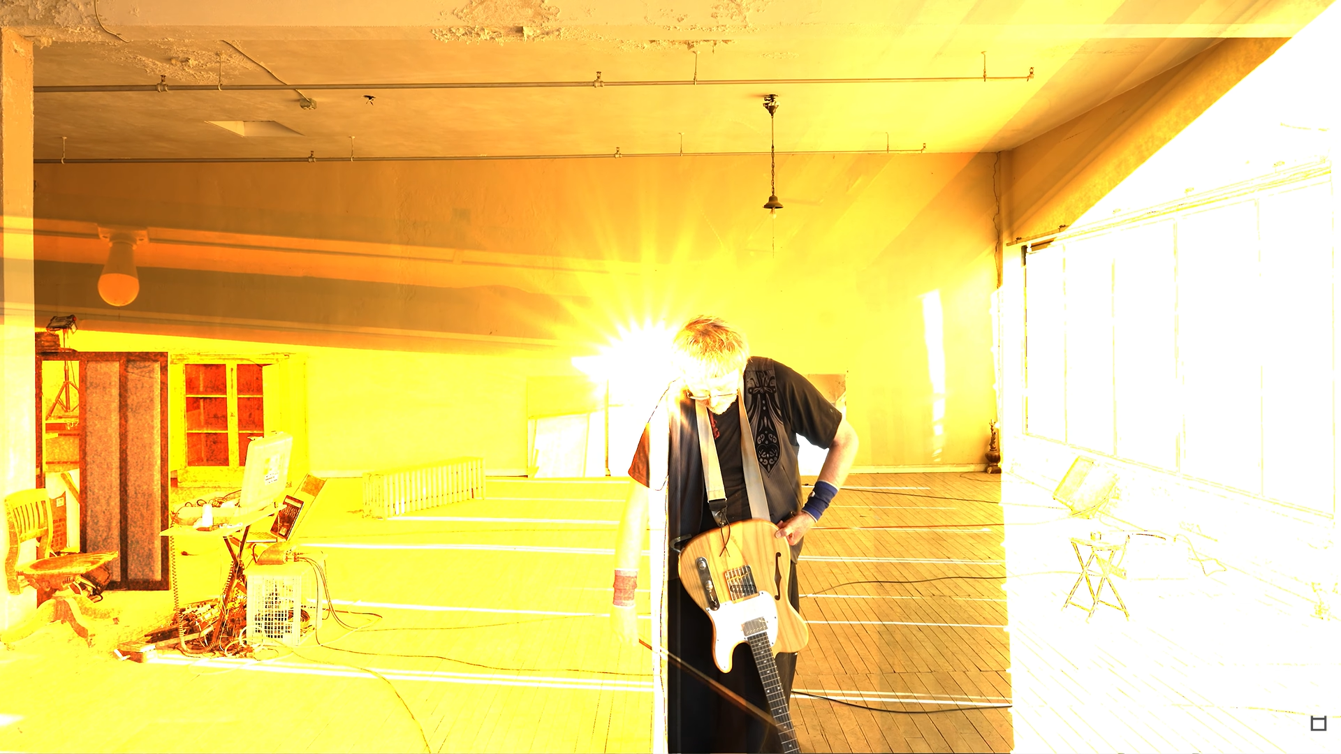 A man with curly hair playing an electric guitar in a brightly lit room, with sunlight streaming in through large windows on the right side of the image.