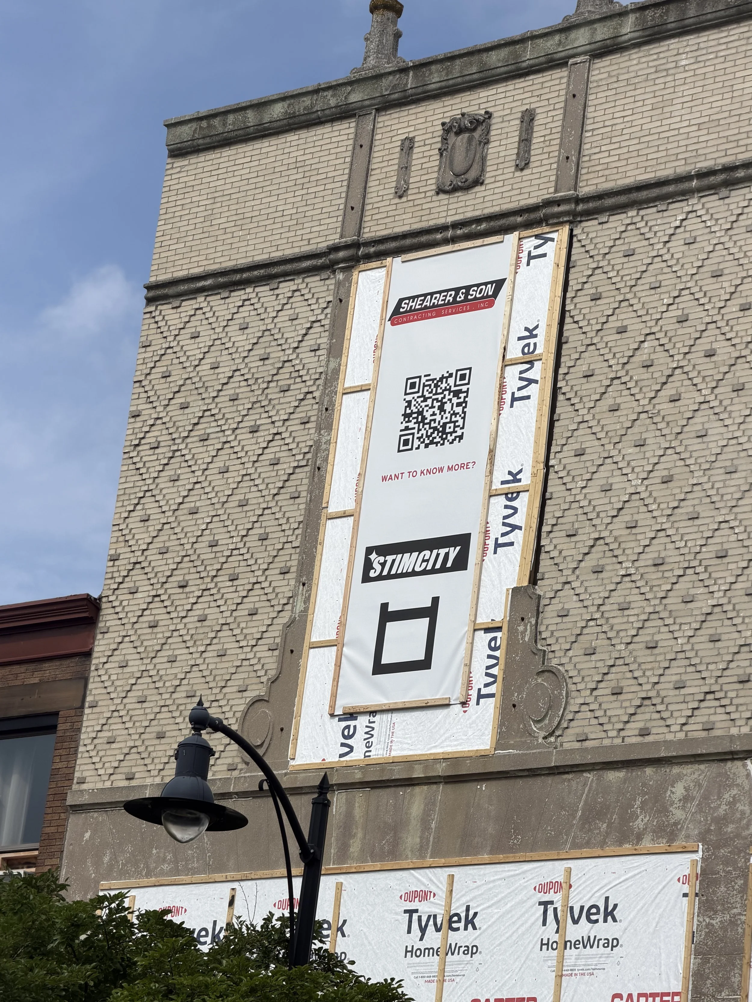 A building under renovation with a large banner advertising Shearer & Son contracting services, featuring a QR code, on a brick wall. The banner is partly covered by Tyvek HomeWrap material and construction framework. A lamppost is visible in the for