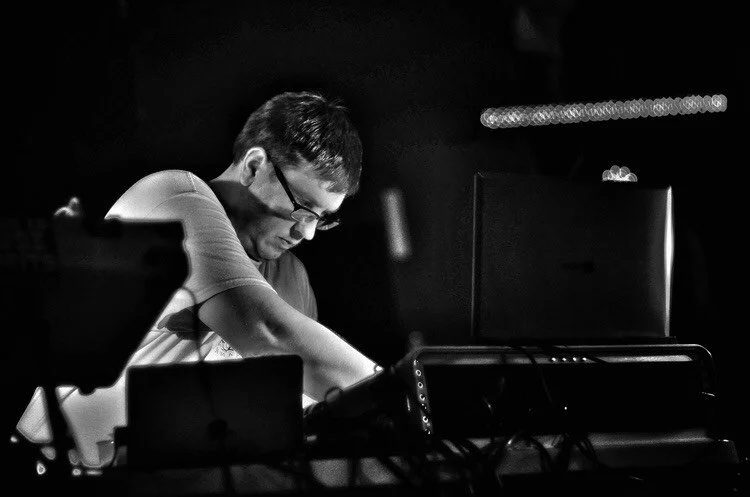 A man wearing glasses sitting at a table with electronic equipment, working on a computer in a dimly lit room.