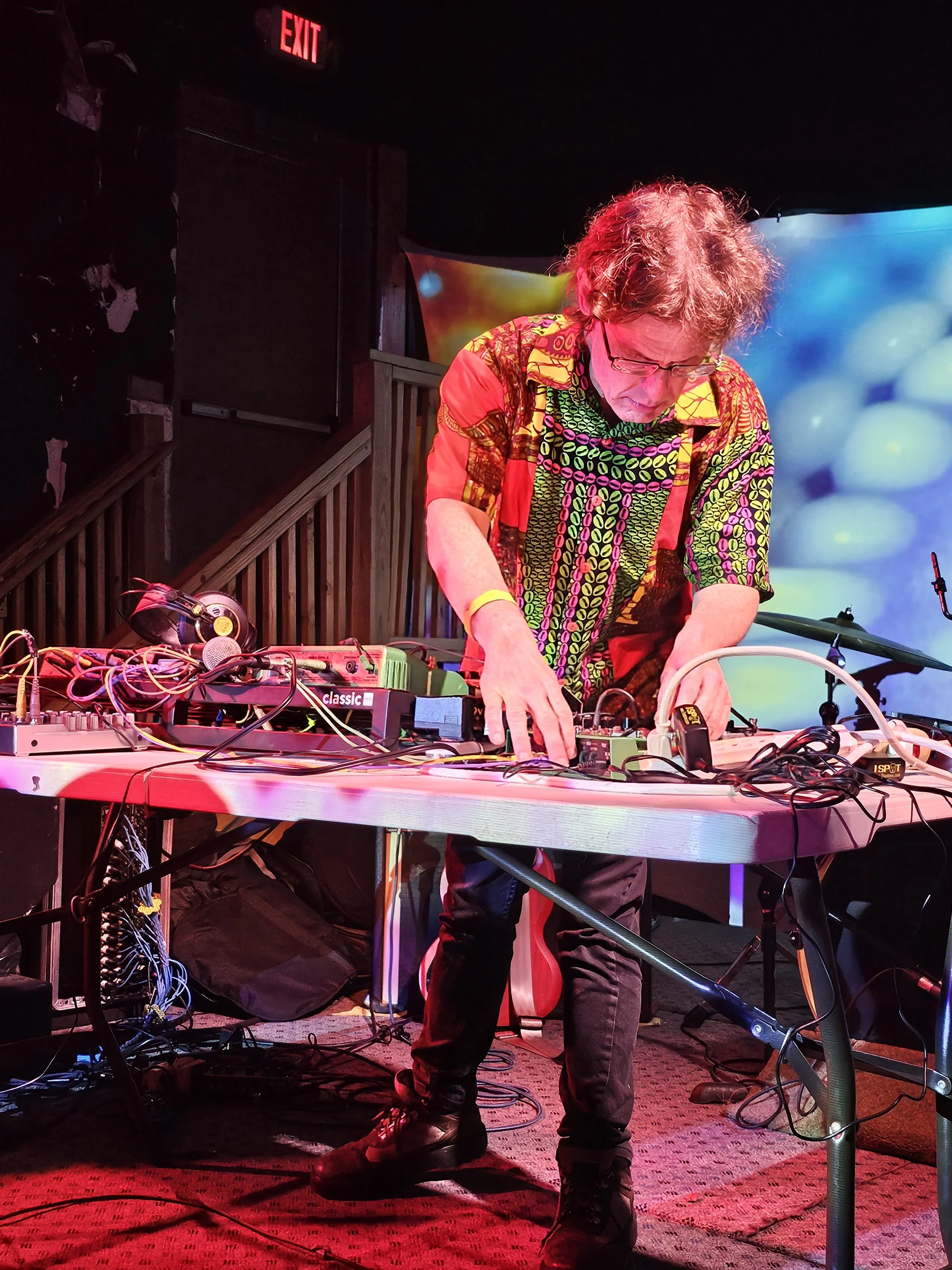 A man with curly hair wearing glasses and a colorful patterned shirt is performing as a DJ on stage, surrounded by music equipment and cables, with a vibrant backdrop and a red exit sign above.