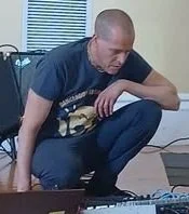 A man crouching on the floor in a room with beige walls and window blinds, wearing a black T-shirt and dark pants.