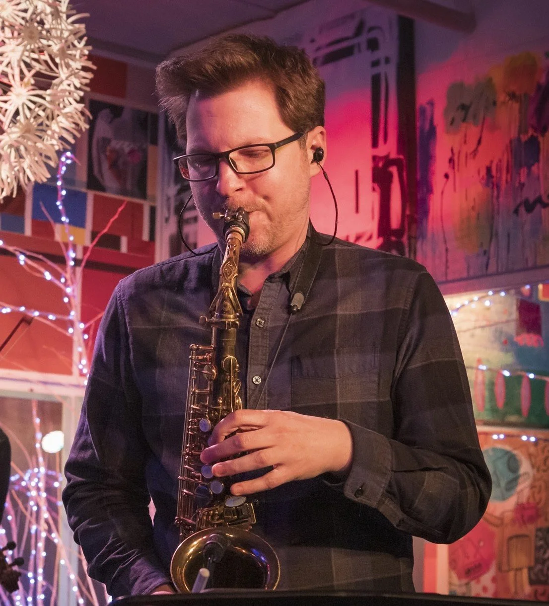 A man wearing glasses playing a saxophone at a colorful indoor venue decorated with string lights and abstract artwork.