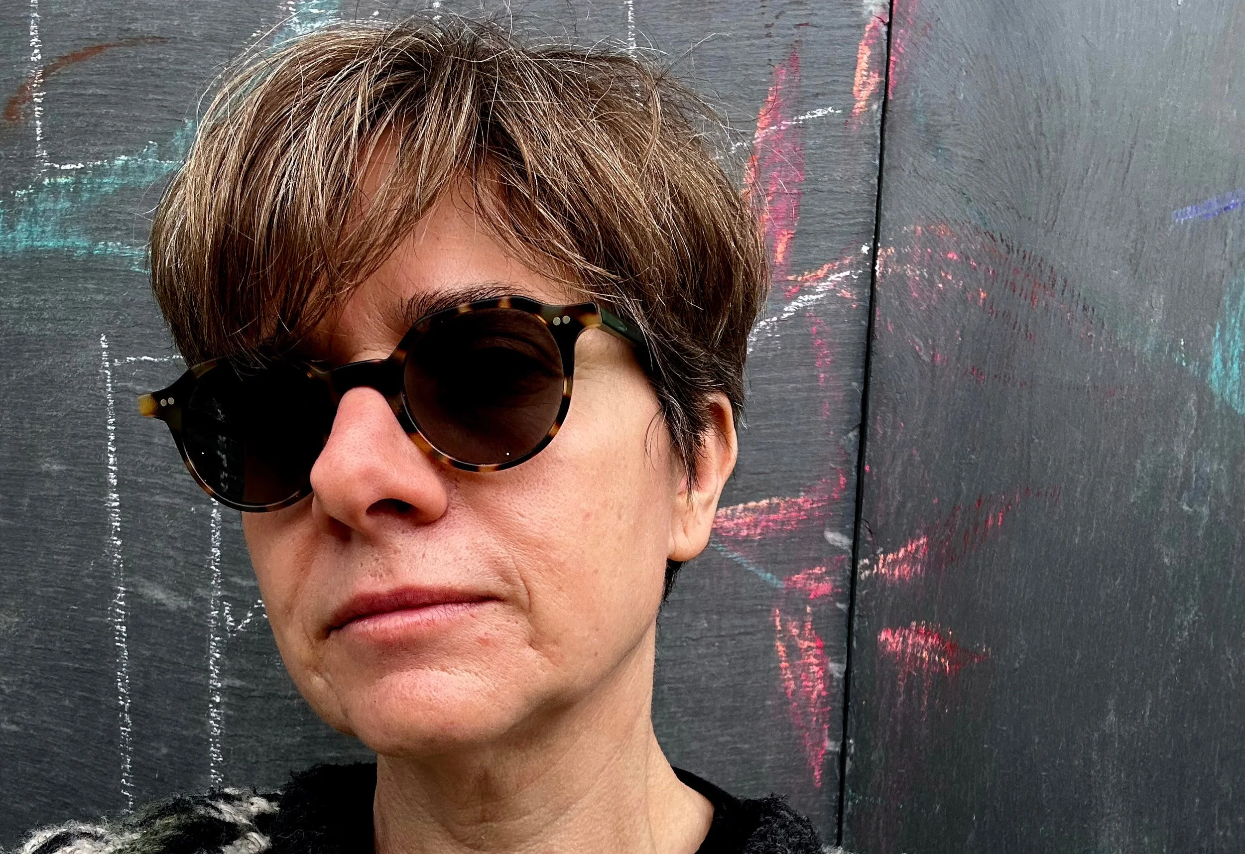 A close-up of a person with short brown hair and round tortoiseshell sunglasses standing in front of a dark painted wall with colorful chalk markings.