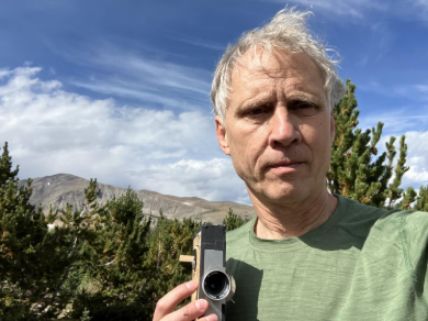 A man with gray hair holding a camera outdoors with trees, mountains, and a blue sky with clouds in the background.