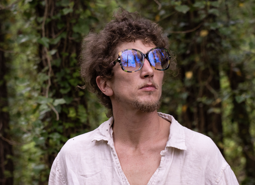 A man with curly hair and glasses standing outdoors in a wooded area.