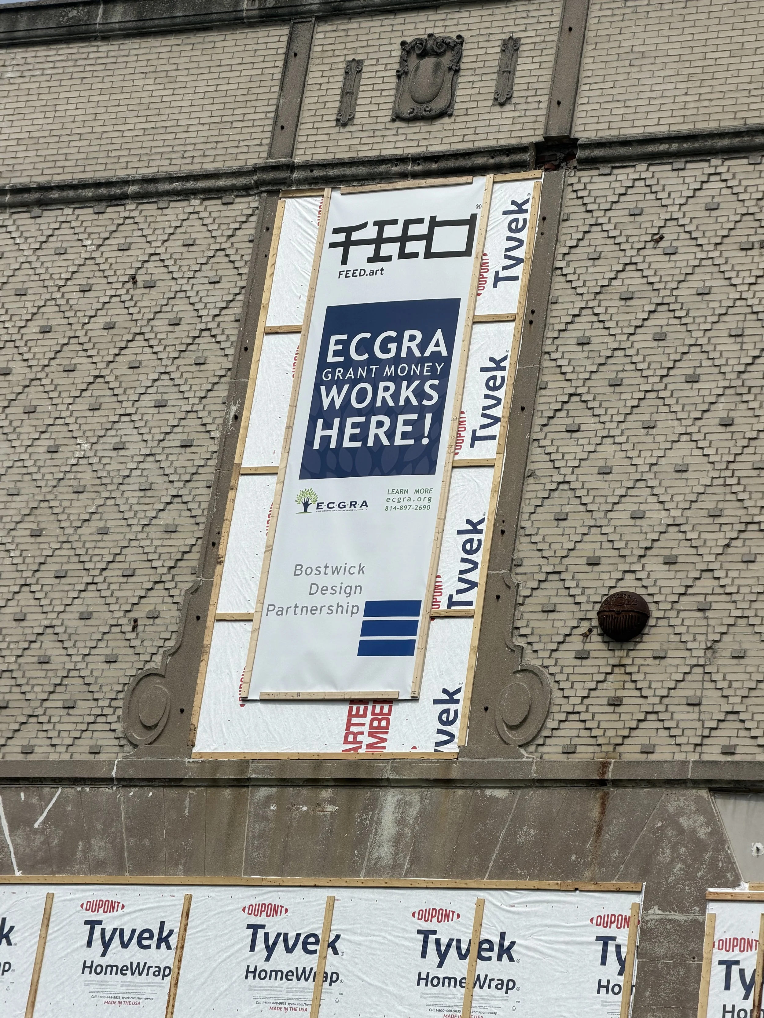 Construction site with a building facade and a large vertical banner that reads 'ECGRA GRANT MONEY WORKS HERE!' and includes contact information, overlaid on brick and stone wall materials.