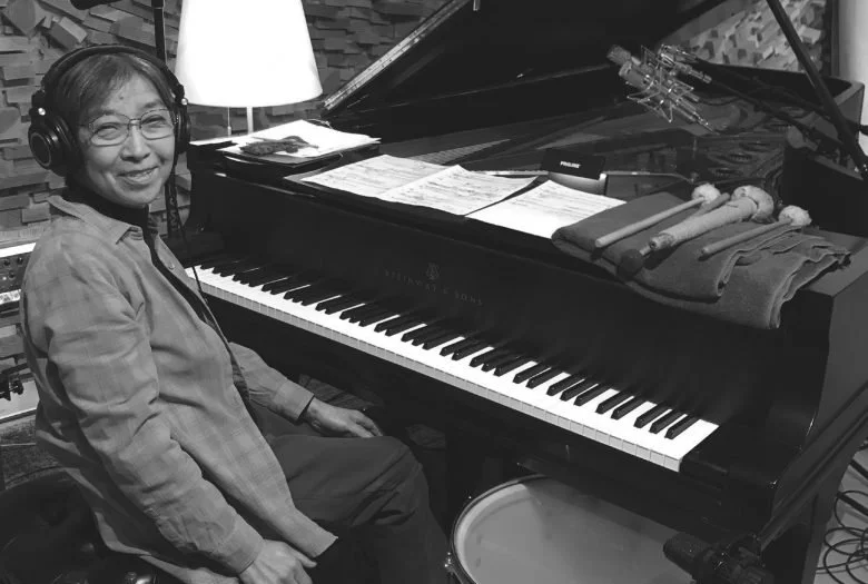A woman sitting at a grand piano in a recording studio, wearing headphones and smiling.