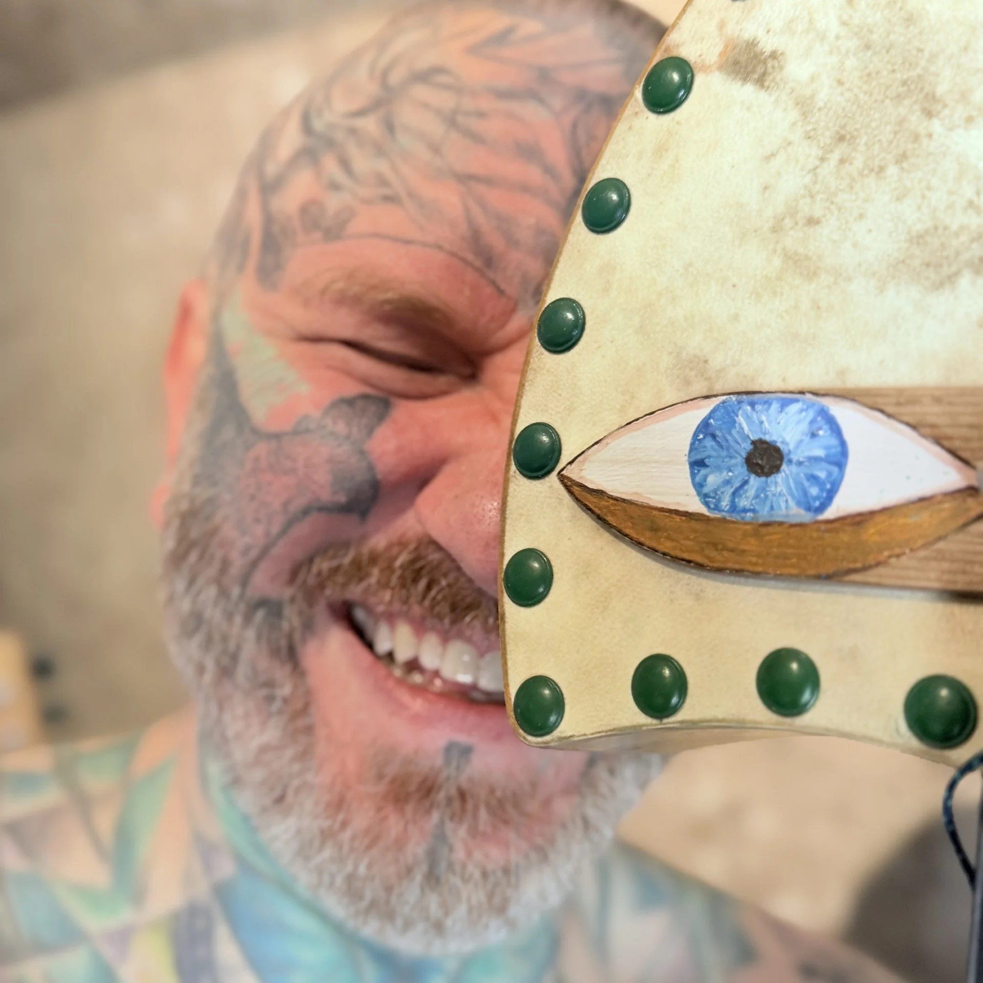 Close-up of a smiling man with colorful tattoos holding a decorated object with blue eye designs and green metallic bumps.