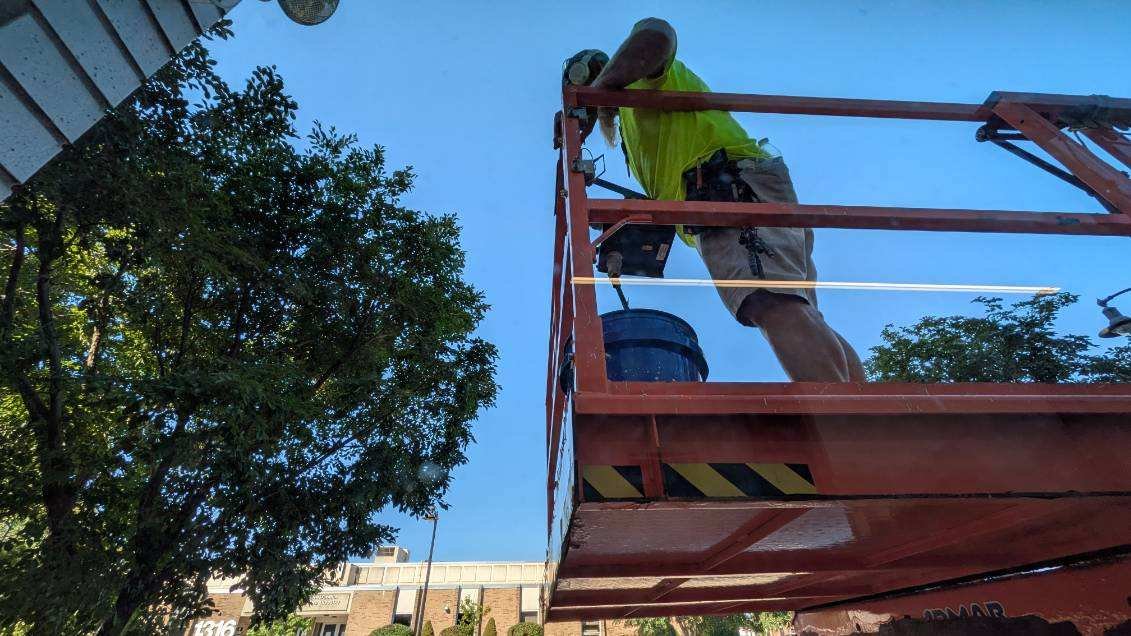 A worker on a red hydraulic lift wearing a bright yellow safety vest and a helmet, working on a high structure, possibly repairing or installing something.