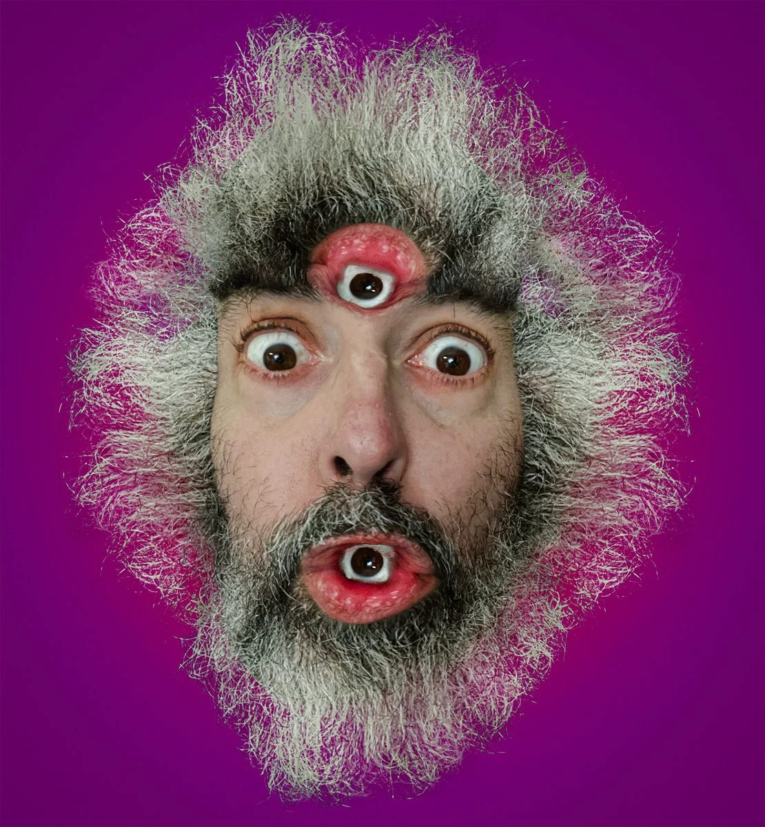 A surreal image of a man's face with gray hair and beard, featuring three eyes. Two eyes are on his face, and one eye is on his forehead amid the gray hair, with the eyes on the forehead and chin resembling eyeballs.