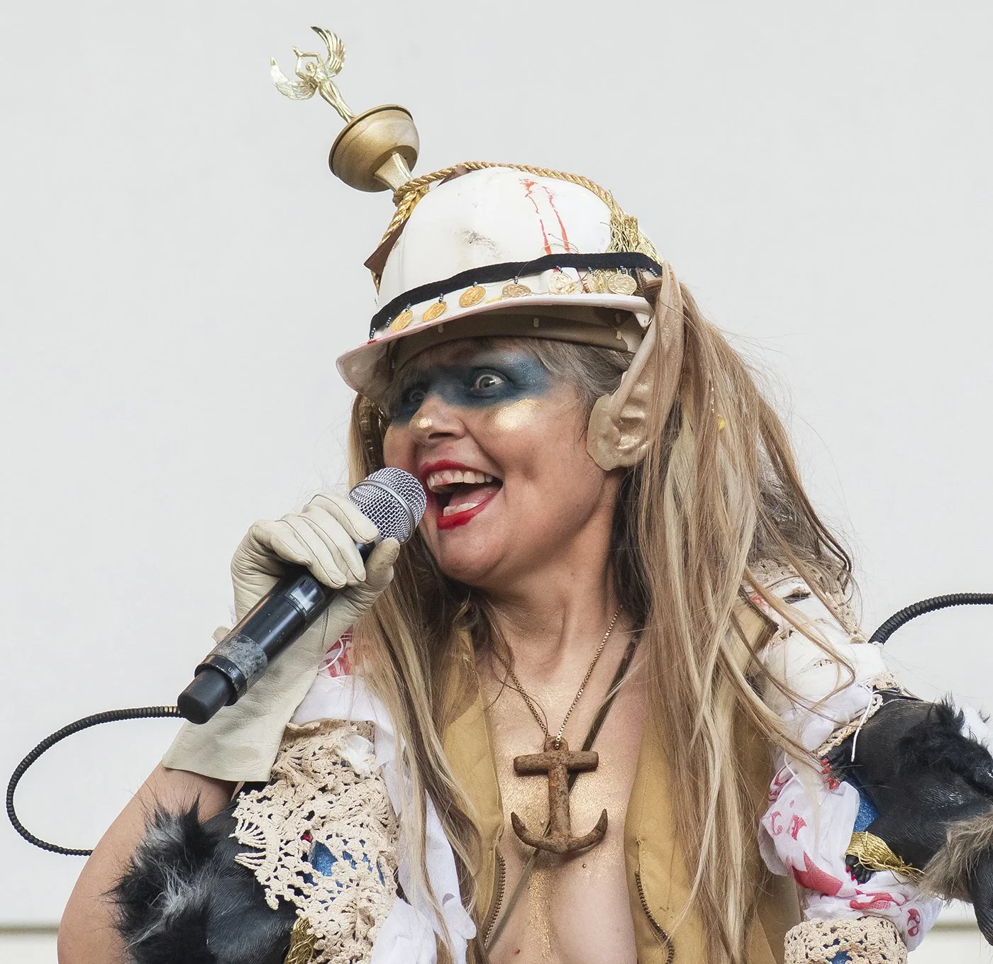 Woman dressed as a pirate singing into a microphone, wearing a hat with an anchor and an anchor necklace, with elf-like ears and elaborate makeup.