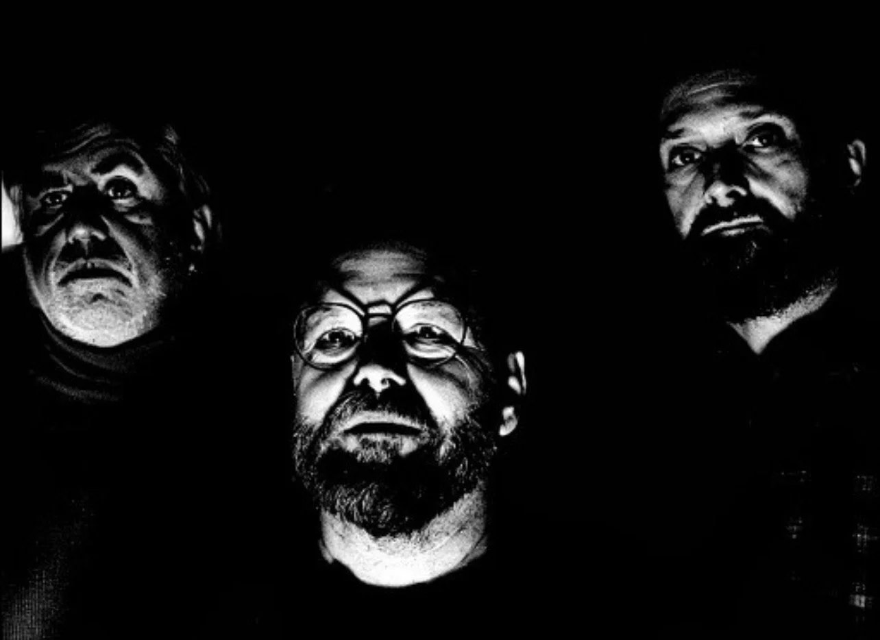 Black and white photo of three men with serious expressions, illuminated from below, against a dark background.