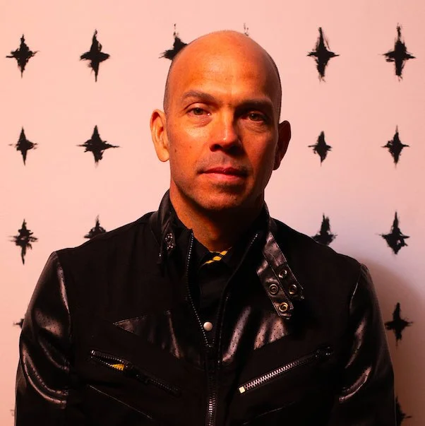 A bald man in a black leather jacket standing in front of a pink wall decorated with black star patterns.