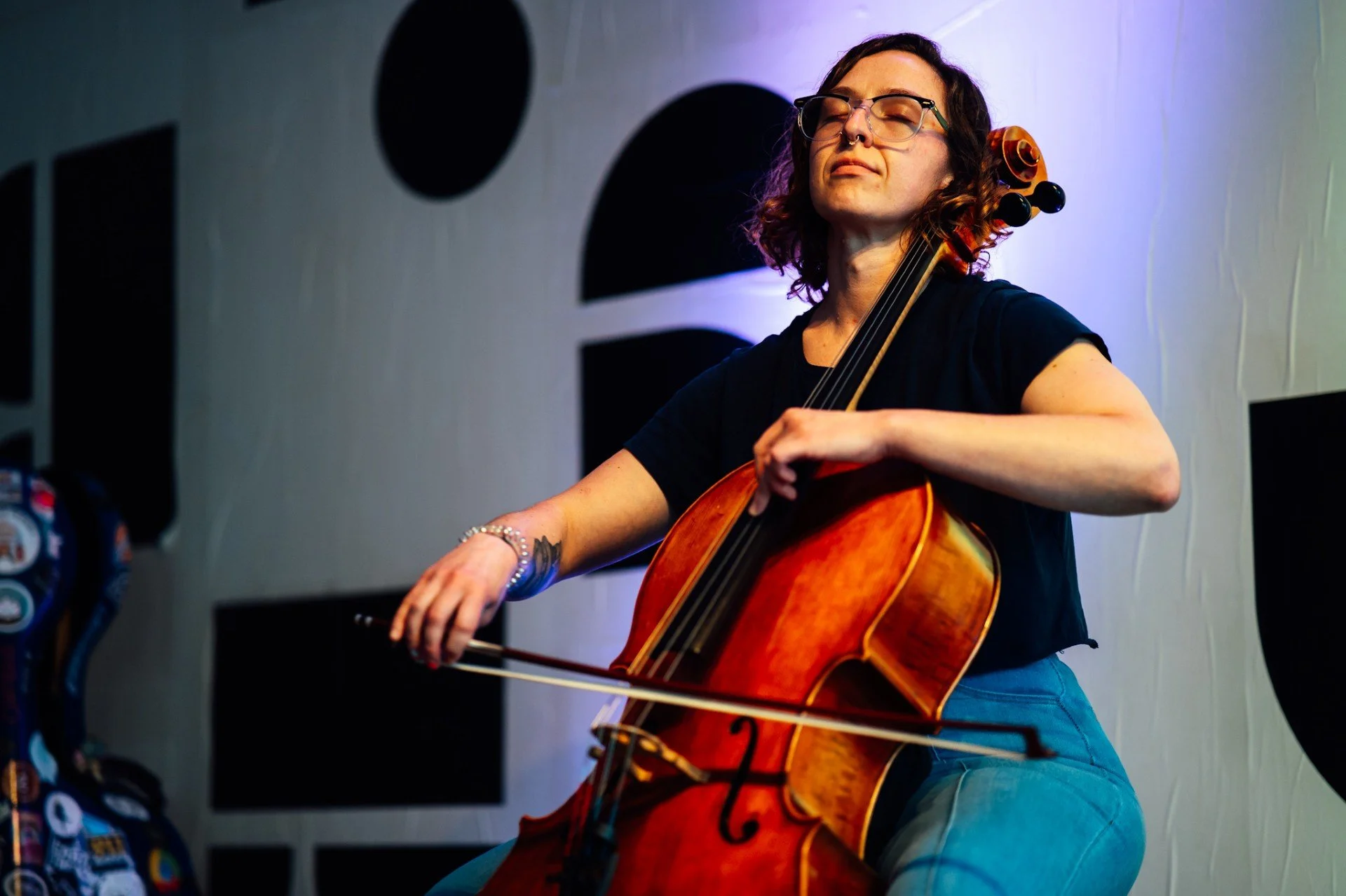 A woman with glasses and curly hair playing a cello with her eyes closed in a room with soundproofing panels.