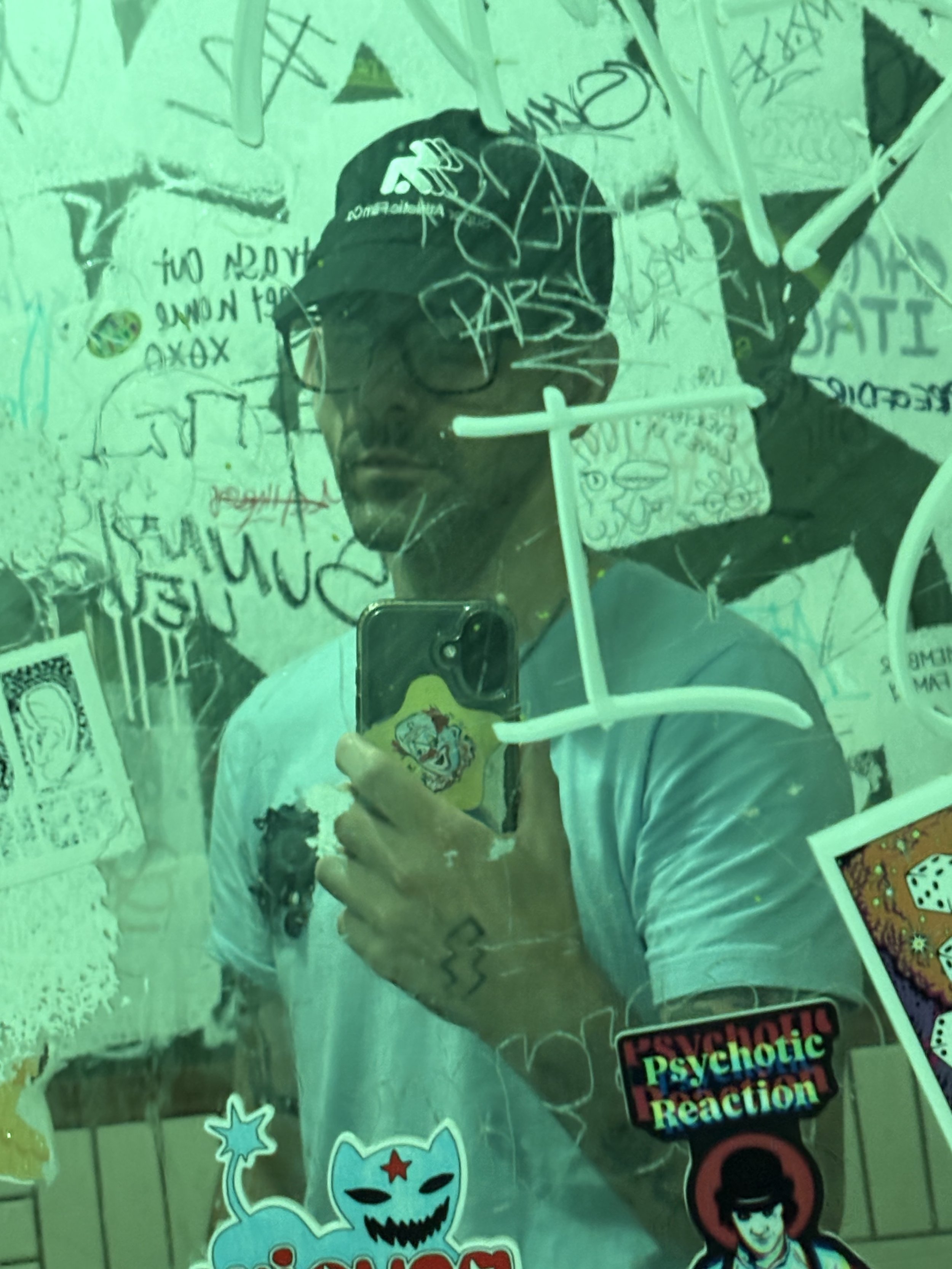 A person wearing sunglasses and a black cap taking a selfie in front of a mirror covered with colorful graffiti and stickers, including a sticker with the words 'psychotic reaction' and a cartoon of a clown.