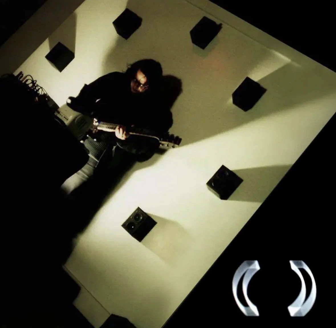 Person playing electric guitar in a room with multiple wall speakers and stage lighting.