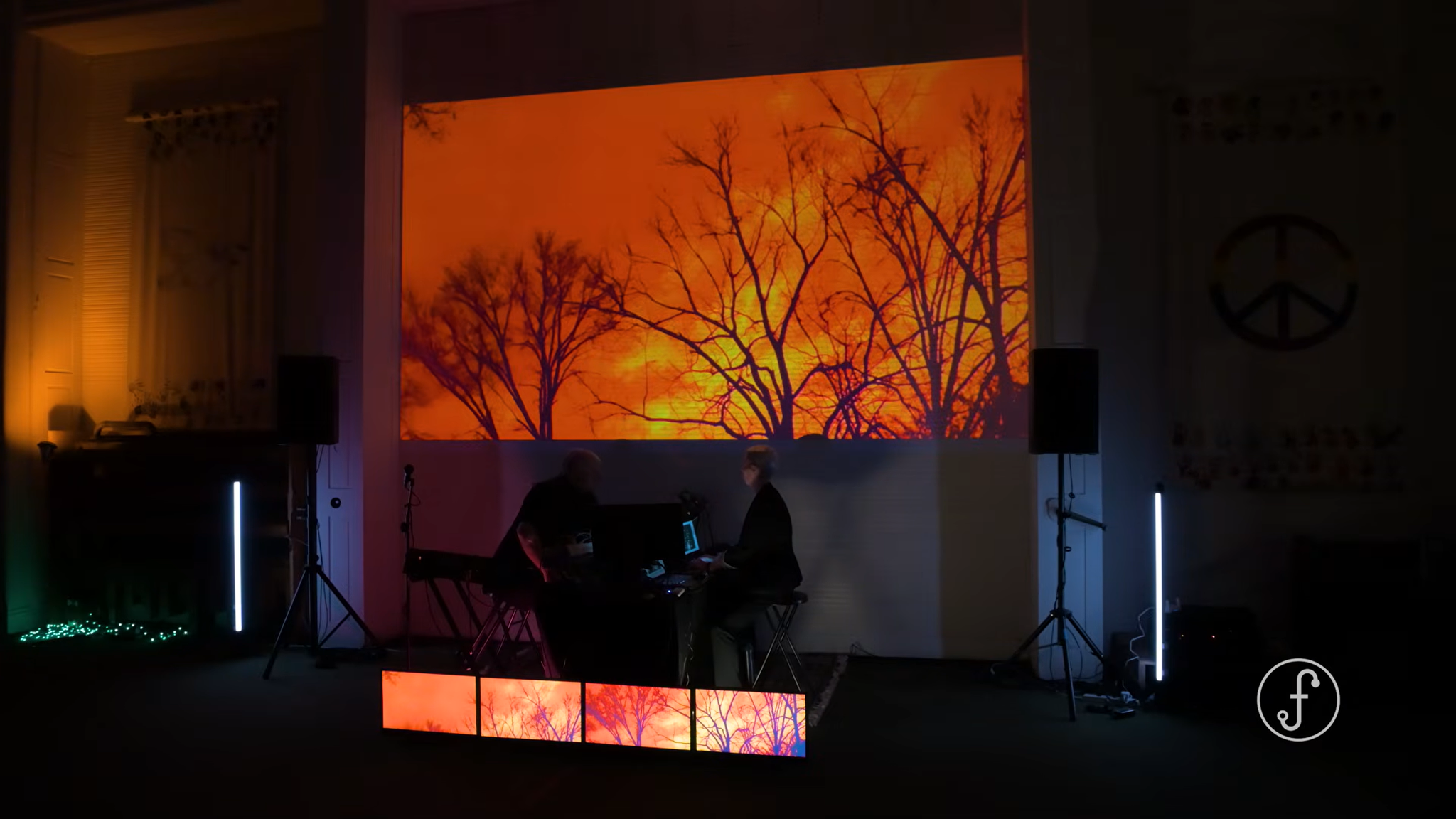 A dark room with a large screen displaying the silhouette of trees against an orange sky. There are two people working at a desk, surrounded by equipment, with vertical light sticks on either side.