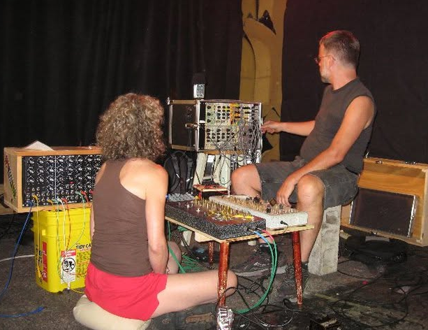 Two people in a music studio working with audio equipment. One is sitting on a small stool, facing a mixing console, with curly hair and wearing a brown tank top and red shorts. The other, a man with glasses, is sitting on a wooden block, adjusting equipment on a rack.