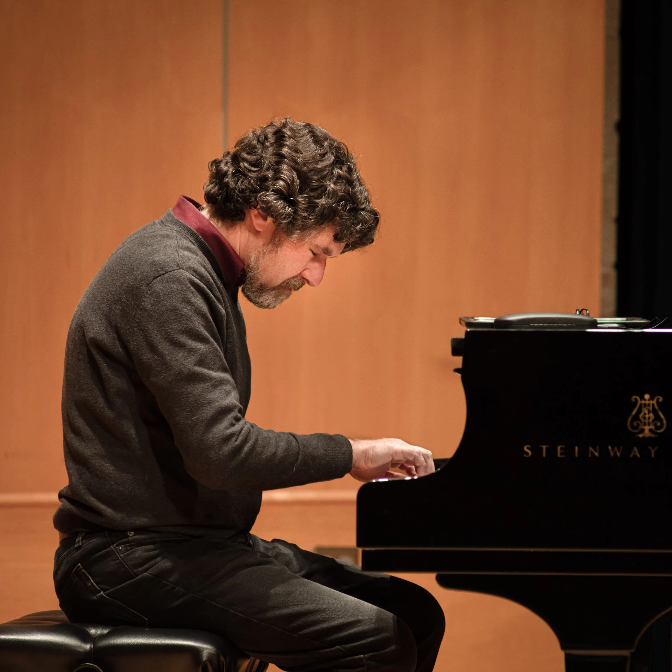 A man with curly brown hair, a beard, and wearing a gray sweater over a maroon shirt, is playing a grand piano on a stage with a wooden background.