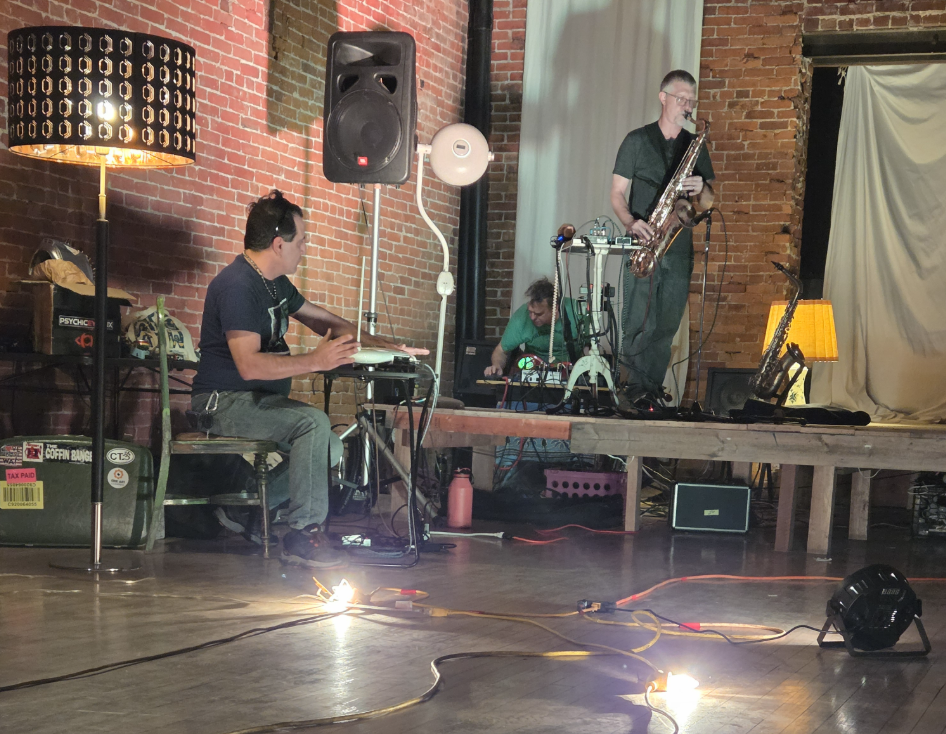 Three musicians performing on a small stage inside a brick-walled venue. One is playing a keyboard, another is operating audio equipment, and the third is playing a saxophone. There is a tall lamp, a speaker, and a small side table with a lamp on it.
