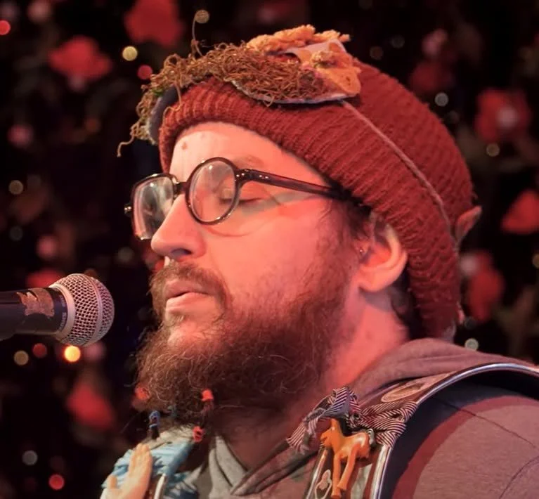 A man with a beard and glasses wearing a red knit cap, singing or speaking into a microphone with a background of red and black lights.