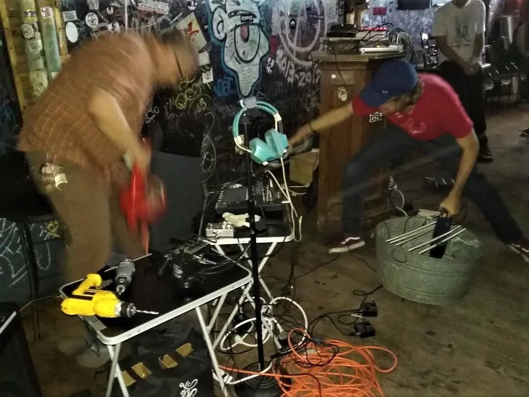 Two men working with electronic equipment and cables in a dimly lit room with graffiti on the wall, a small table with tools, and headphones hanging from a stand.