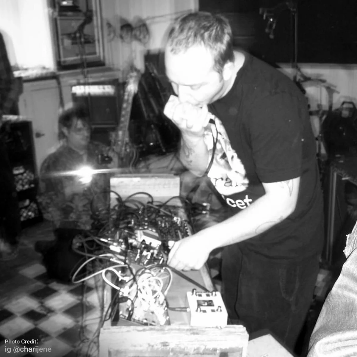 A man with glasses and tattooed arms is performing electronic music with a mixer and cables in a room filled with music equipment, while a woman with glasses sits in the background.
