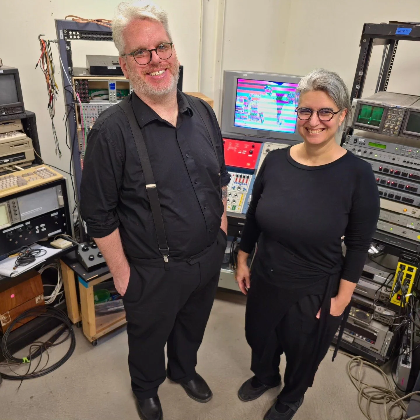 Two smiling people, a man with white hair and glasses and a woman with gray hair and glasses, standing in a room filled with electronic equipment and monitors, likely a control or editing room.