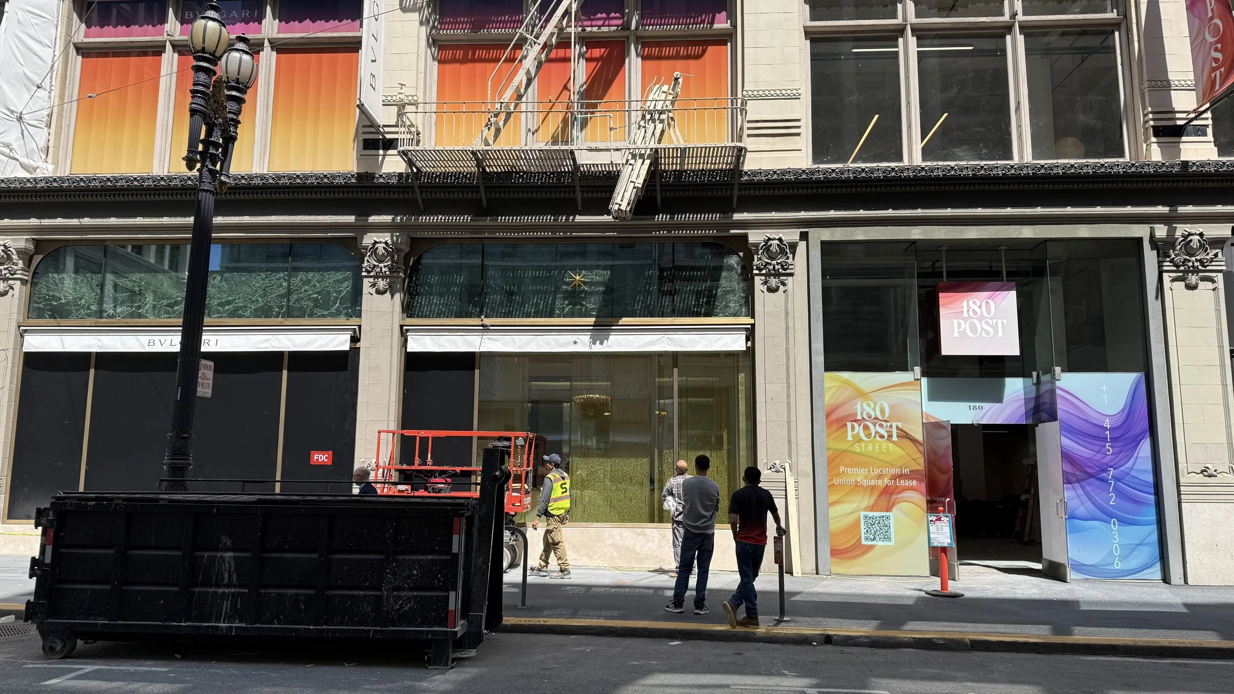 Construction workers standing in front of a storefront with a colorful poster that reads '180 POST' and 'Premier Location in Union Square for Lease'; debris and equipment are on the sidewalk, with a black dumpster and a red scaffolding.