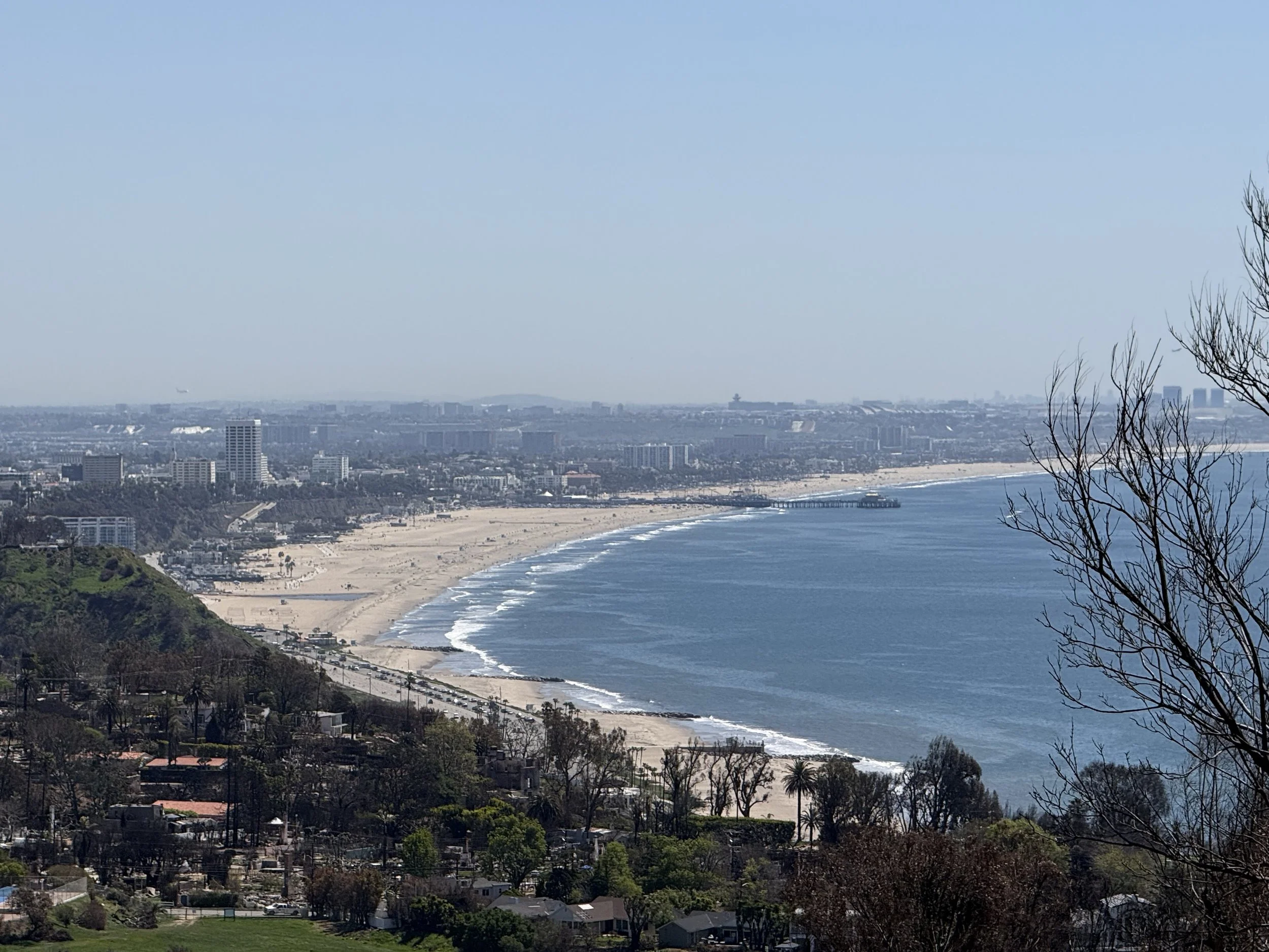 Panoramic view of a beach and ocean with a cityscape background, including a long stretch of sandy beach, a pier, and various buildings in the distance.