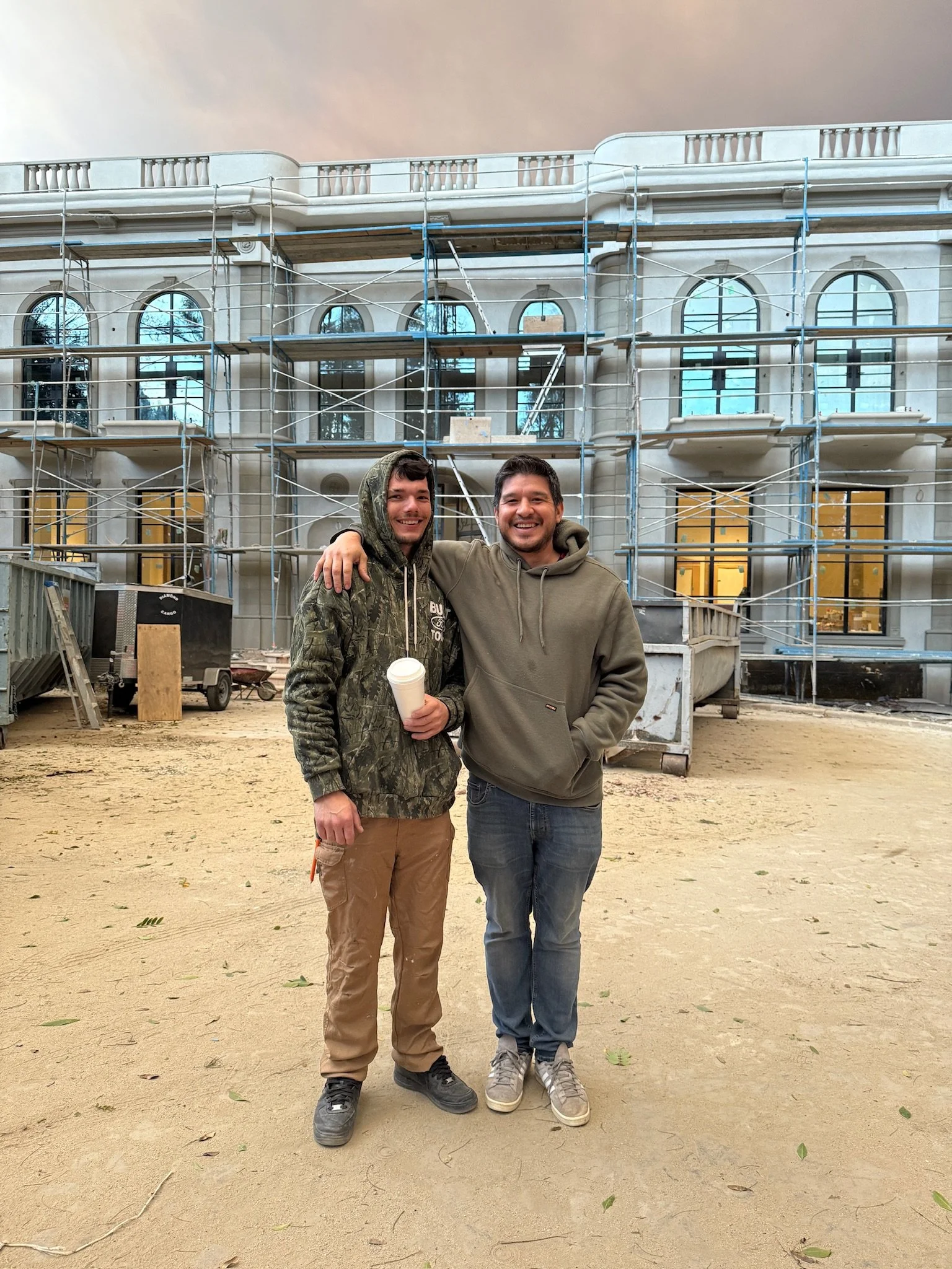 Two smiling men standing in front of a building under construction with scaffolding. One man is wearing a camouflage hoodie and the other a gray hoodie, holding a coffee cup.