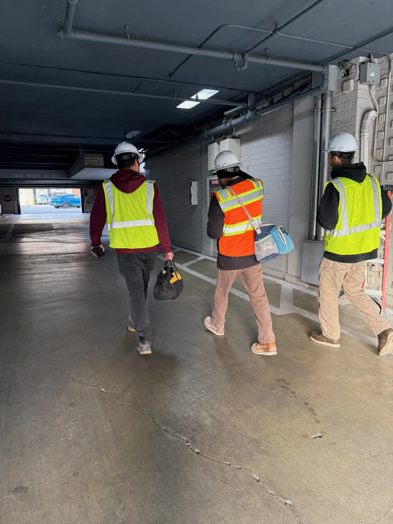 Three construction workers walking through a parking garage, wearing safety vests and helmets, carrying bags and equipment. showing that safety is very important