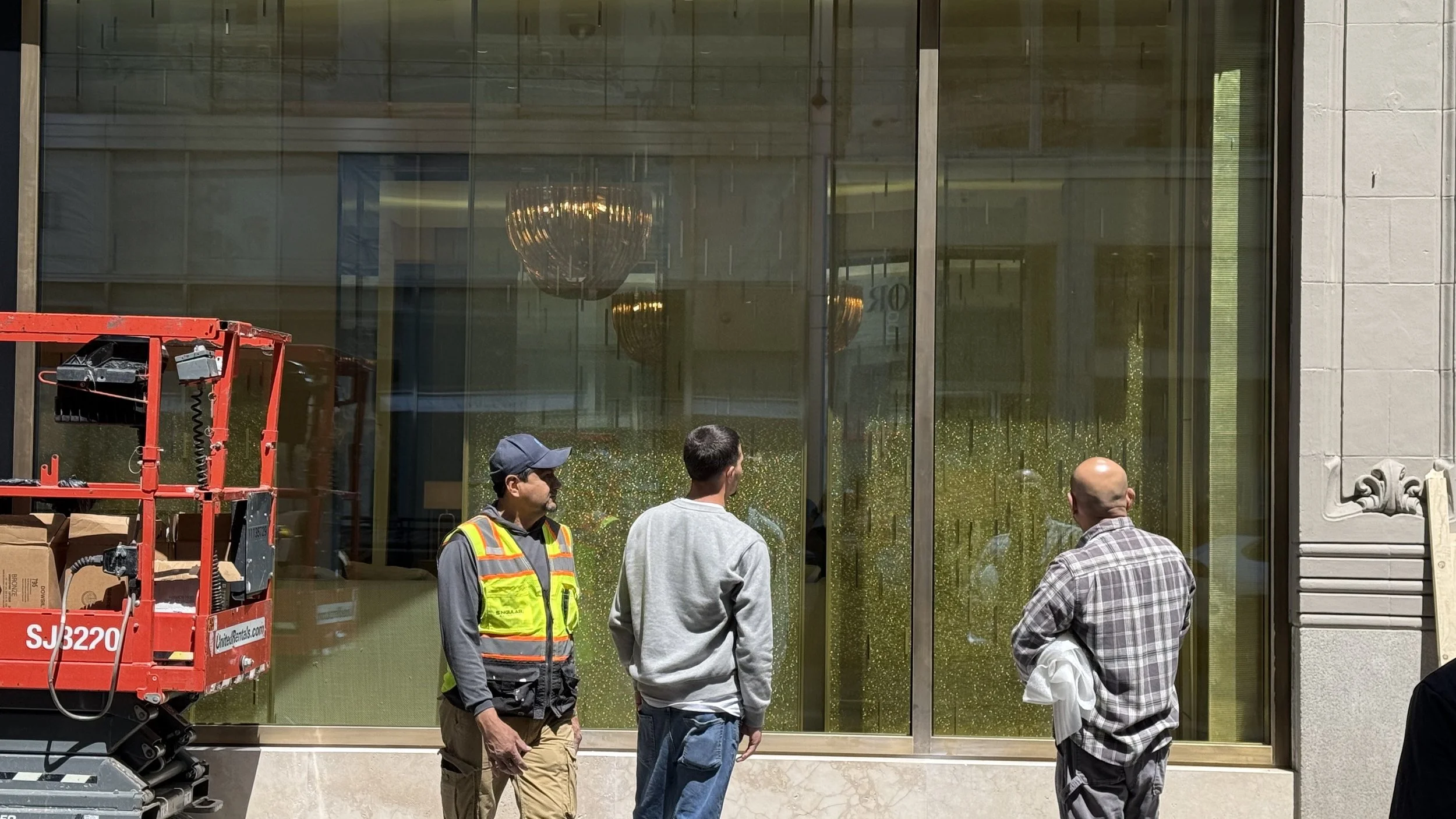 Three men outside a building with large glass windows. One man is in a safety vest and cap, another has a gray hooded sweatshirt, and the third is in a plaid shirt. There is a red lift or scaffold on the left side, and chandeliers and patterned green