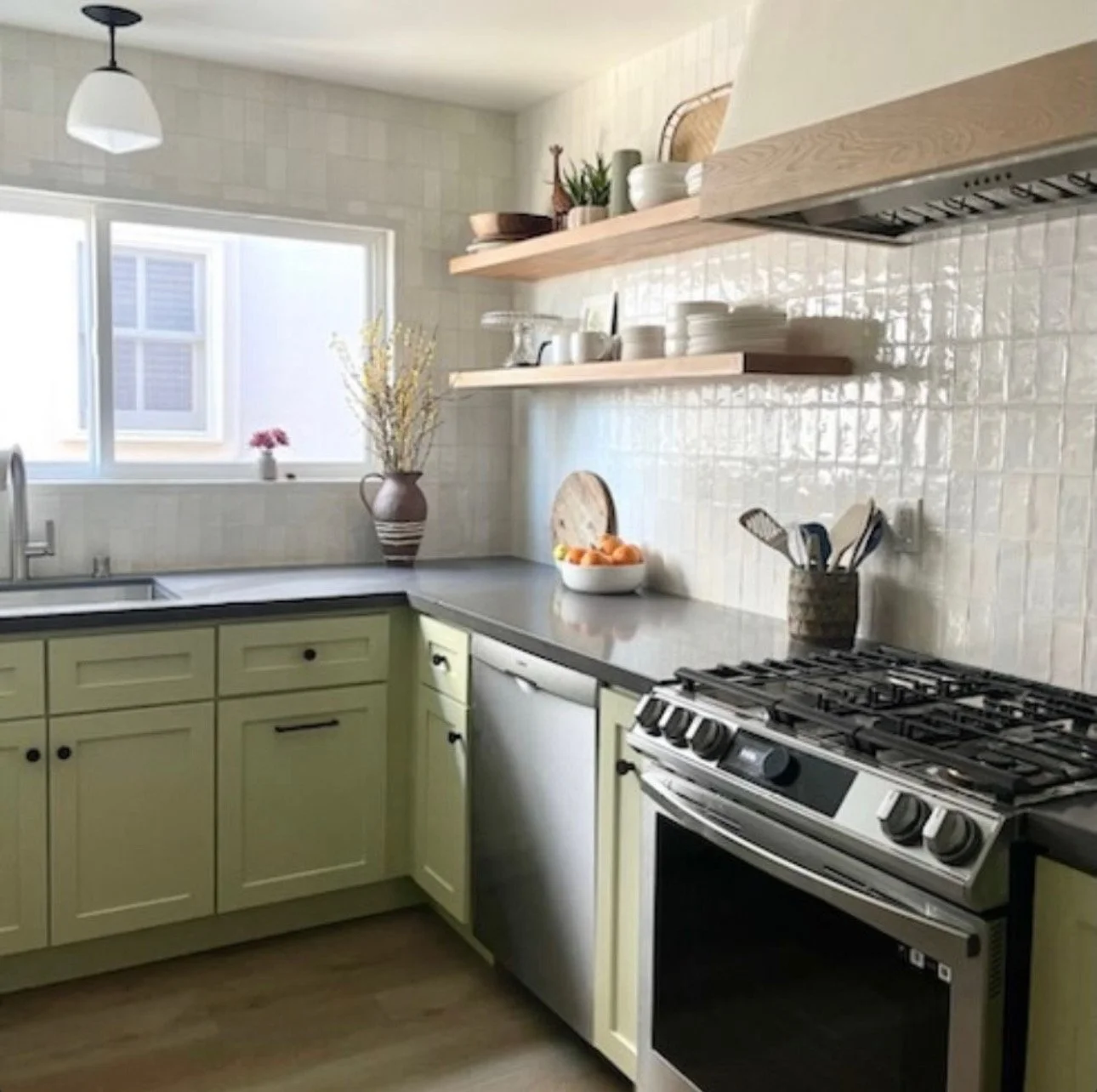 A modern kitchen with light green cabinets, stainless steel appliances, and two wooden open shelves with dishes and decorative items, illuminated by natural light from a window.